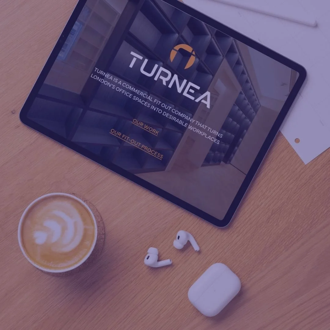 A tablet displaying the TURNEA company website, a cup of latte with latte art, a pair of wireless earbuds, and their charging case on a wooden desk.