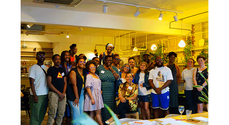 A group of diverse people gathered in a warmly lit modern cafe or restaurant for a group photo, smiling and standing close together.