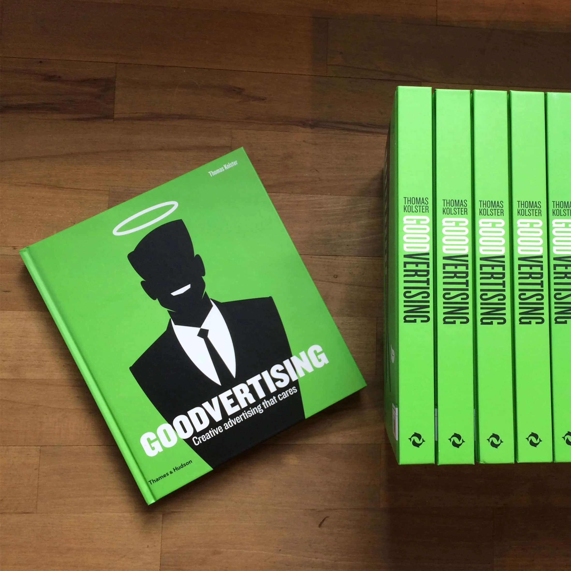 A green book titled 'Good Advertising' lying on a wooden surface with a cartoon illustration of a person in a suit on cover, and a row of matching green books titled 'Good Vertising' standing upright next to it.