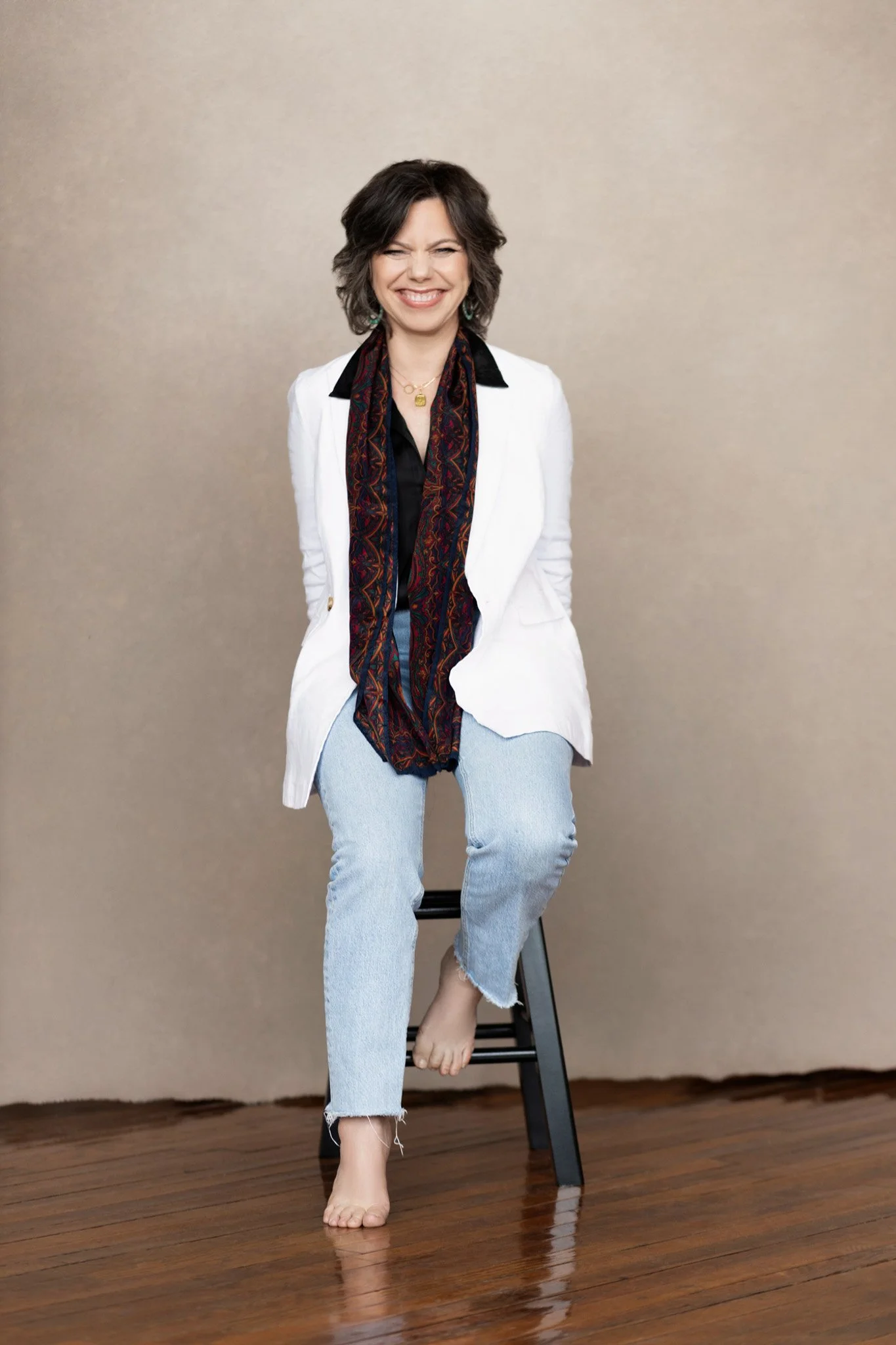 Brunette woman in a white blazer and jeans seated barefoot on a stool, laughing, against a taupe backdrop