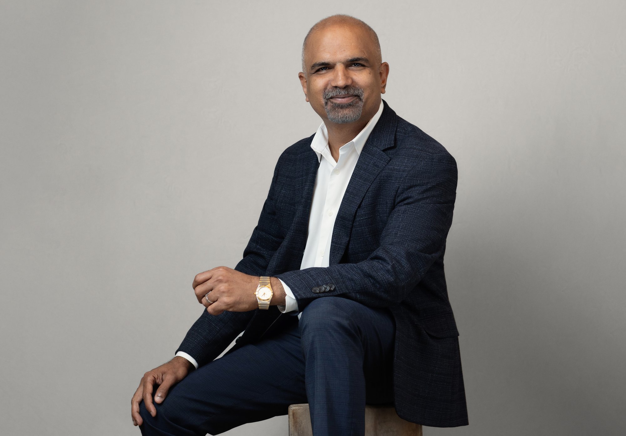 Corporate headshot of a man seated on a stool wearing a navy textured blazer over a white shirt, smiling against a neutral gray backdrop.