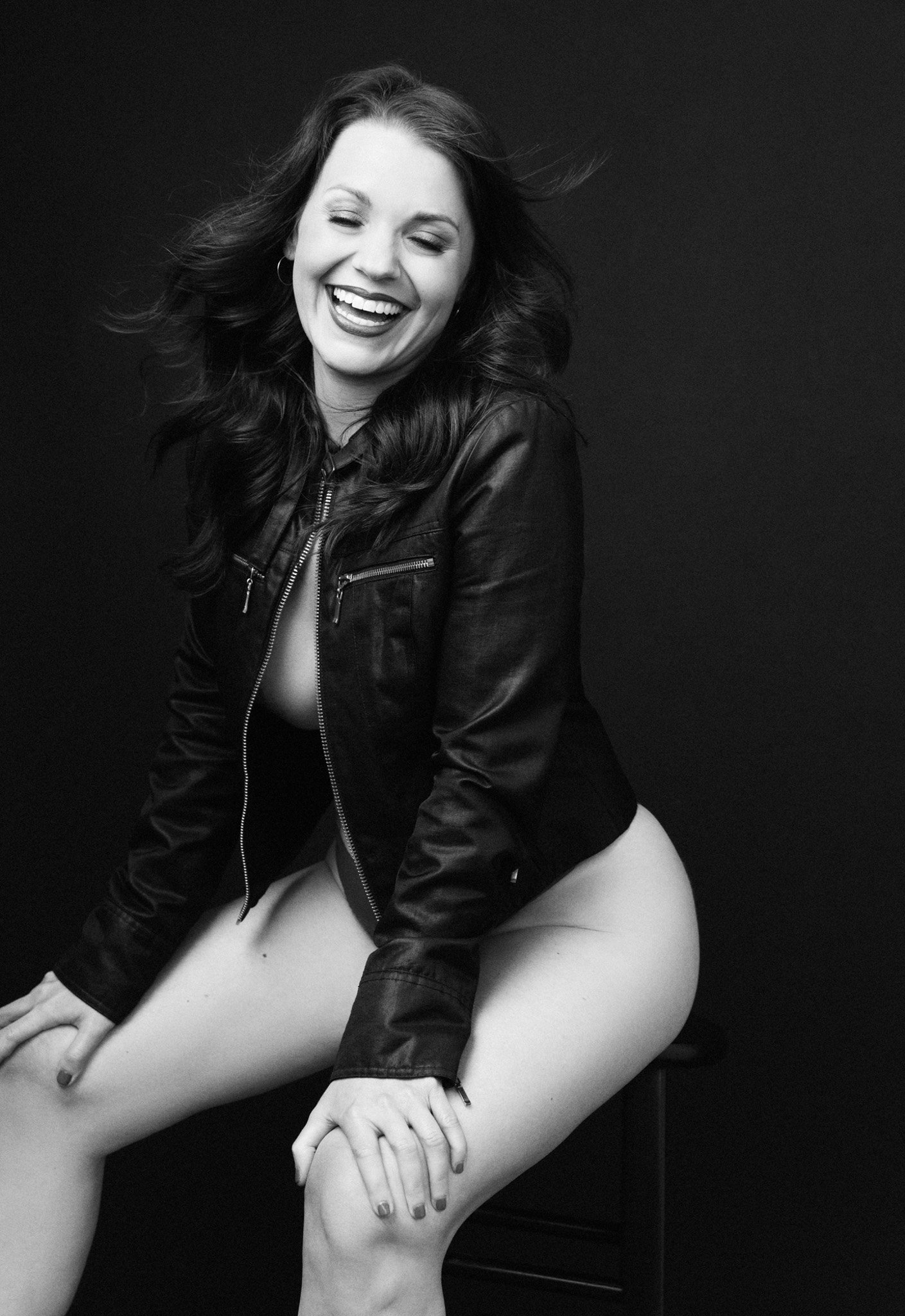 Brunette woman laughing joyfully in an open leather jacket, seated on a stool, black and white — Nashville boudoir photography