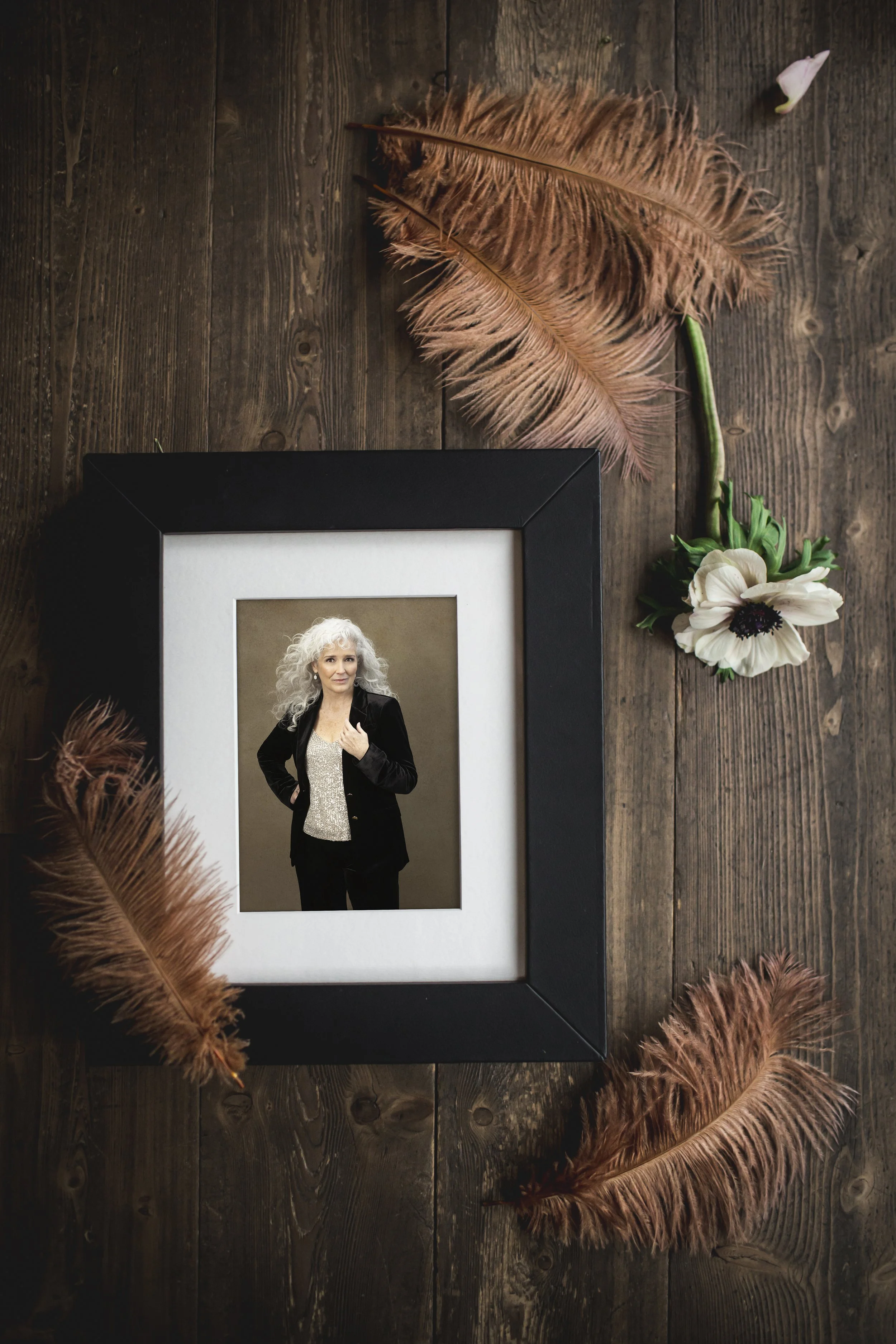 A matted portrait in a thick black frame resting on a dark wood surface.The portrait features a smiling woman with long, curly silver hair wearing a black velvet blazer over a shimmering silver top.