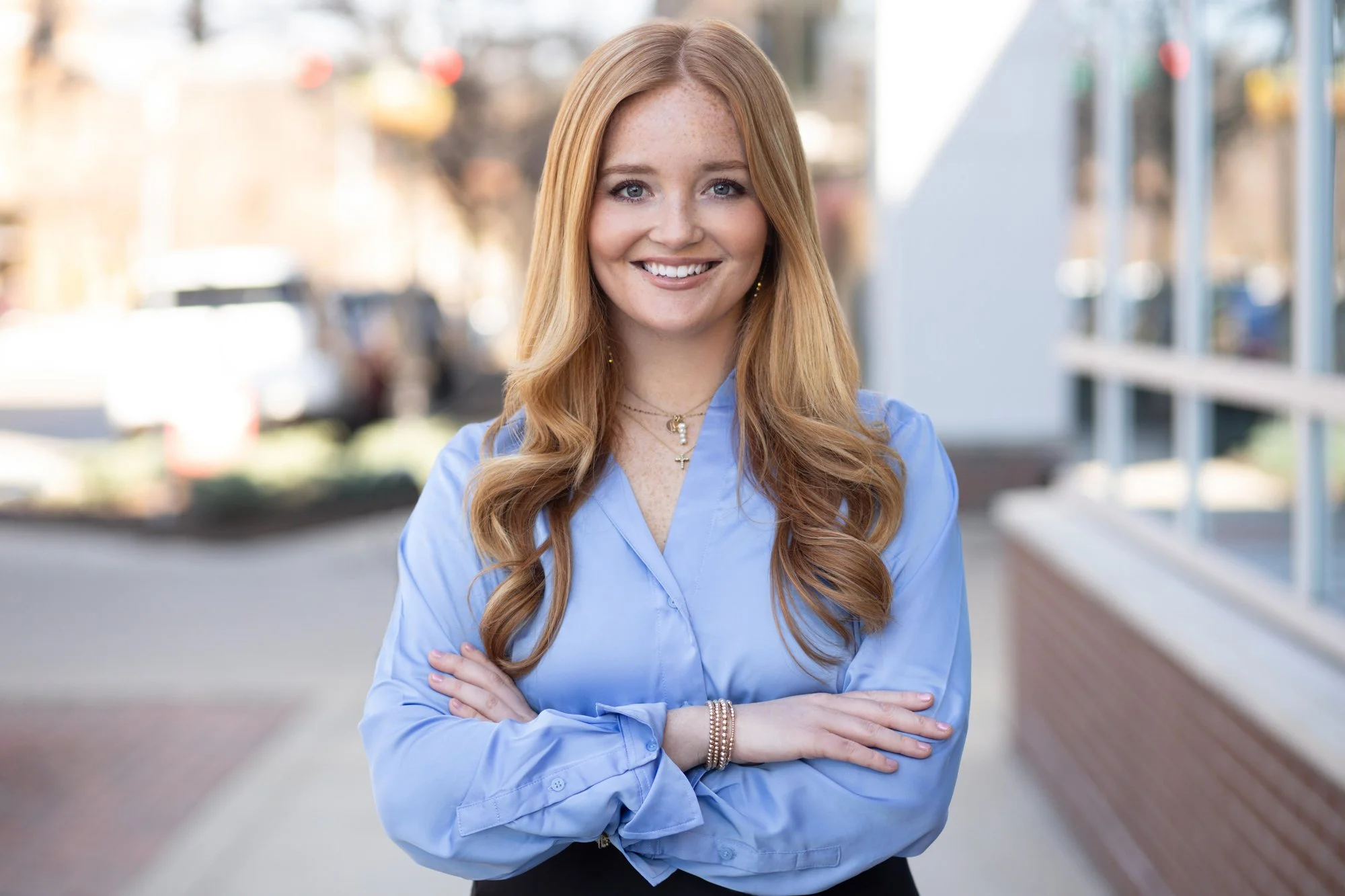 Outdoor corporate headshot of a smiling woman with long strawberry-blonde hair and arms crossed wearing a light blue blouse, on a downtown sidewalk.