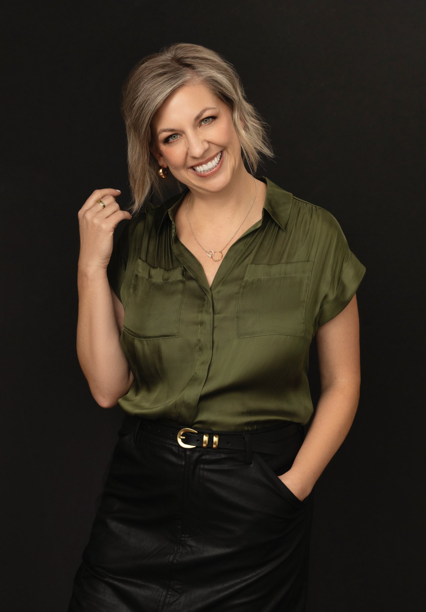  Friendly branding portrait of a blonde woman in her 40s on a black backdrop in an olive green top with hand in pocket and hand in hair. 