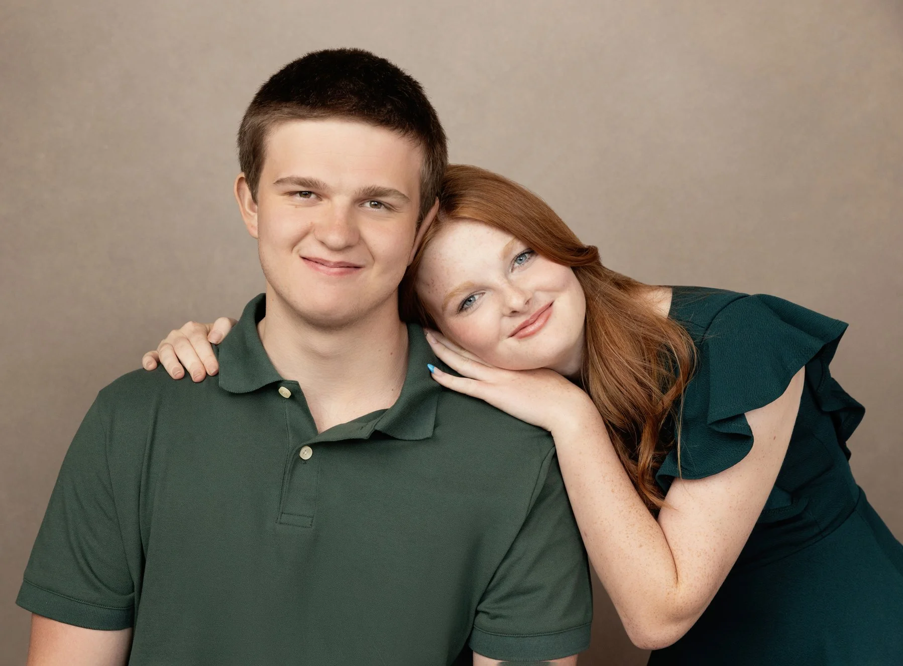  Teenage boy smiling as a red haired girl rests her head on his shoulder, capturing a sweet sibling moment in a family photography session. 