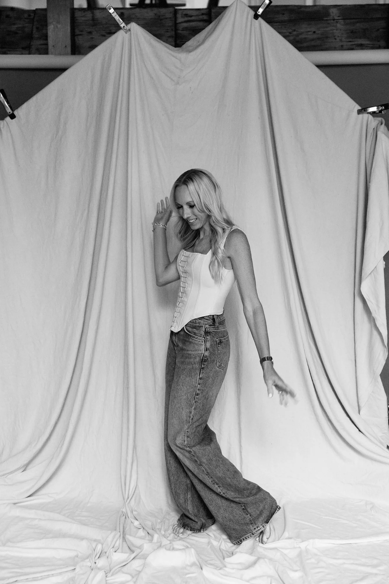  Black and White image of a Woman in her 40s dressed in ivory corset and jeans photographed in Judith Hill Photography’s Nashville Studio on a painter’s tan drop cloth that is draped. 