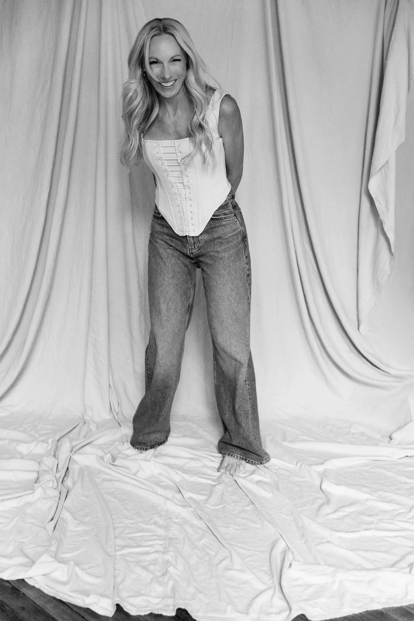  Black and White image of a Woman in her 40s dressed in ivory corset and jeans photographed in Judith Hill Photography’s Nashville Studio on a painter’s tan drop cloth that is draped. 