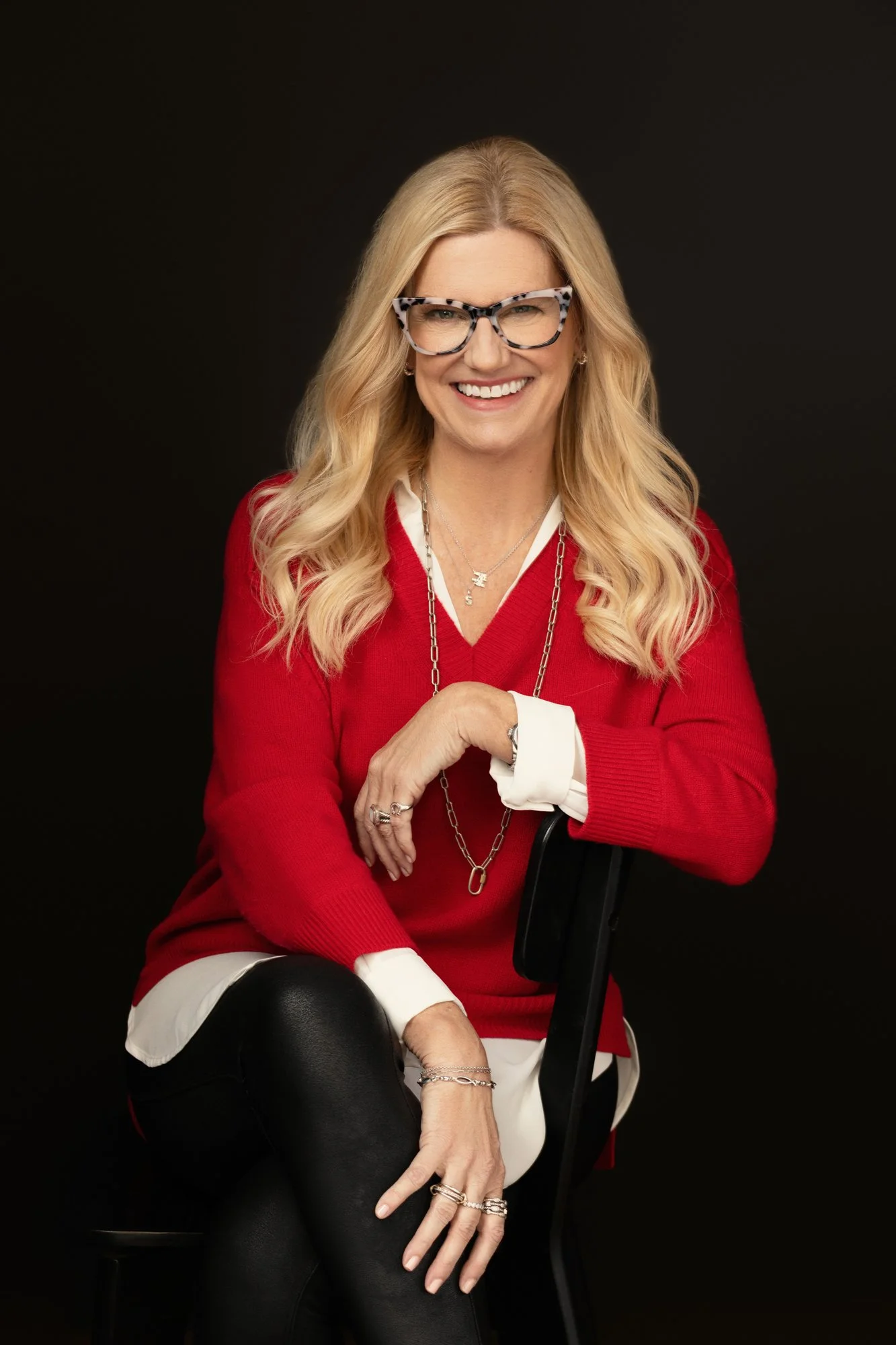  Smiling woman in a red sweater and cat-eye glasses seated on a chair against a black backdrop, Nashville personal branding photography 