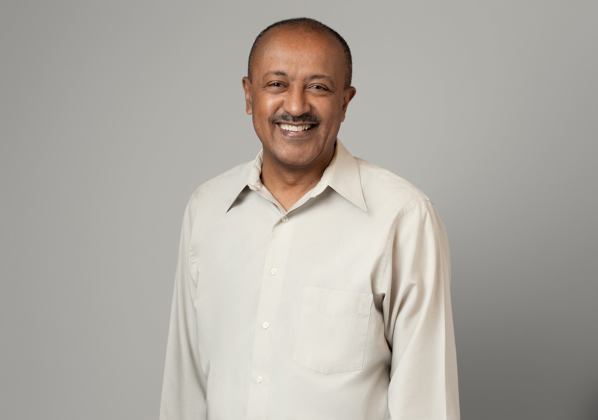 Corporate headshot of a smiling man with a mustache wearing a cream long-sleeve button-up shirt, standing against a light gray background.