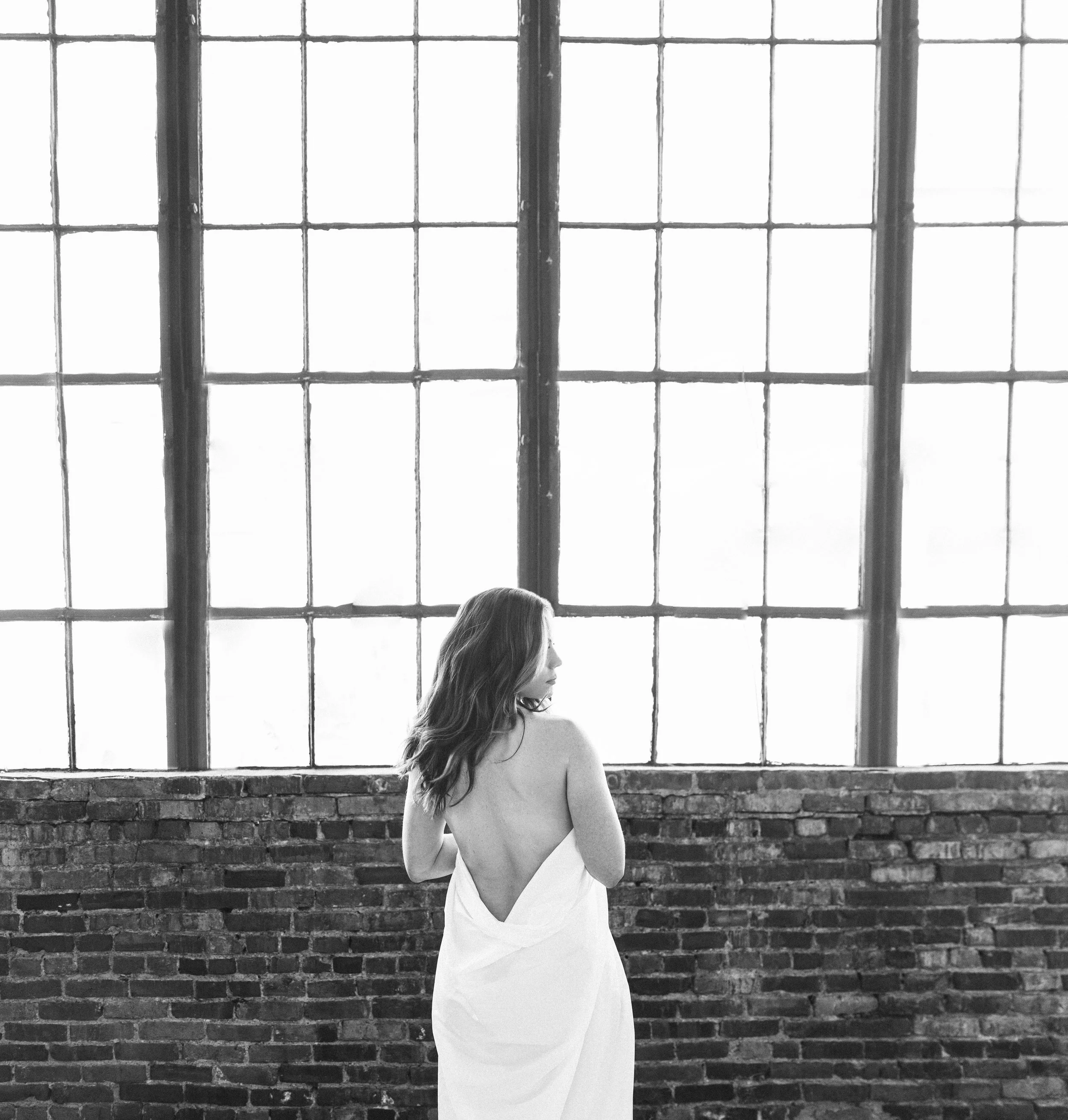 Woman wrapped in a white sheet facing tall factory windows, bare back and brick wall, black and white — Nashville boudoir