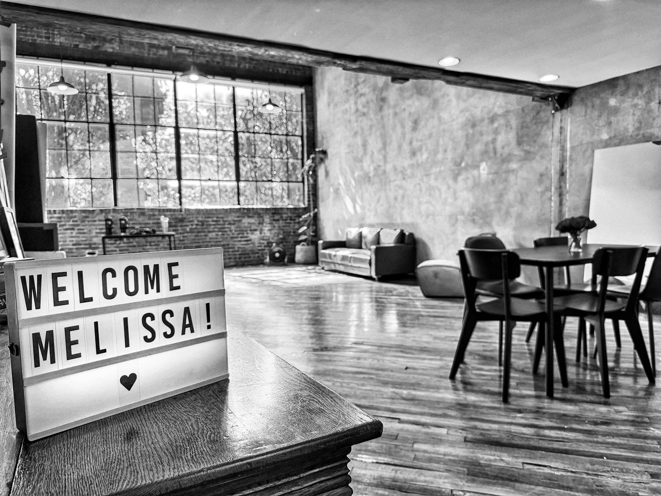 Lightbox sign on a wooden table reads "WELCOME MELISSA!" with a heart symbol. The sign sits inside a bright studio space prepared for a women’s portrait photography session.