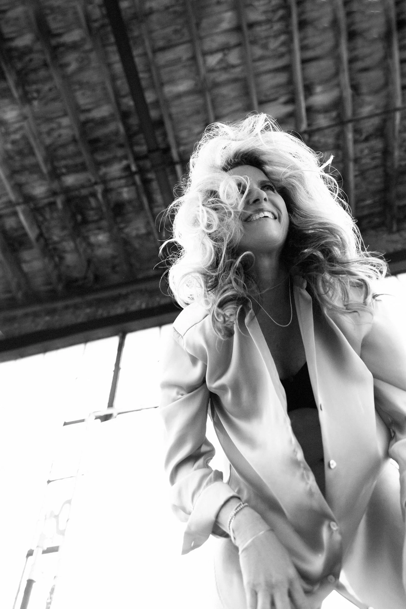 Blonde woman laughing with voluminous curls in a satin blazer, sunlit warehouse, black and white — Nashville boudoir
