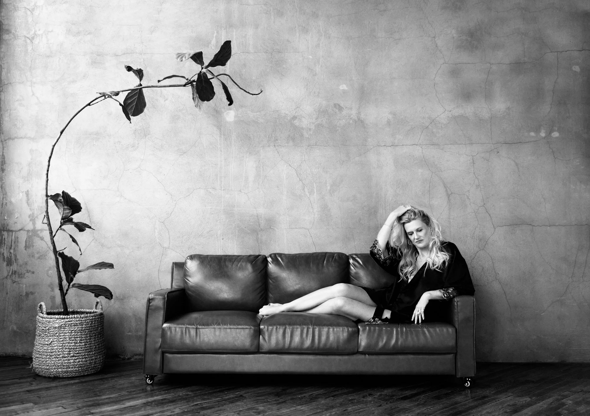 Blonde woman reclines barefoot on a dark leather couch beside a tall potted plant in a minimalist studio. The relaxed pose reflects the calm, reflective atmosphere of a women’s portrait photography session.