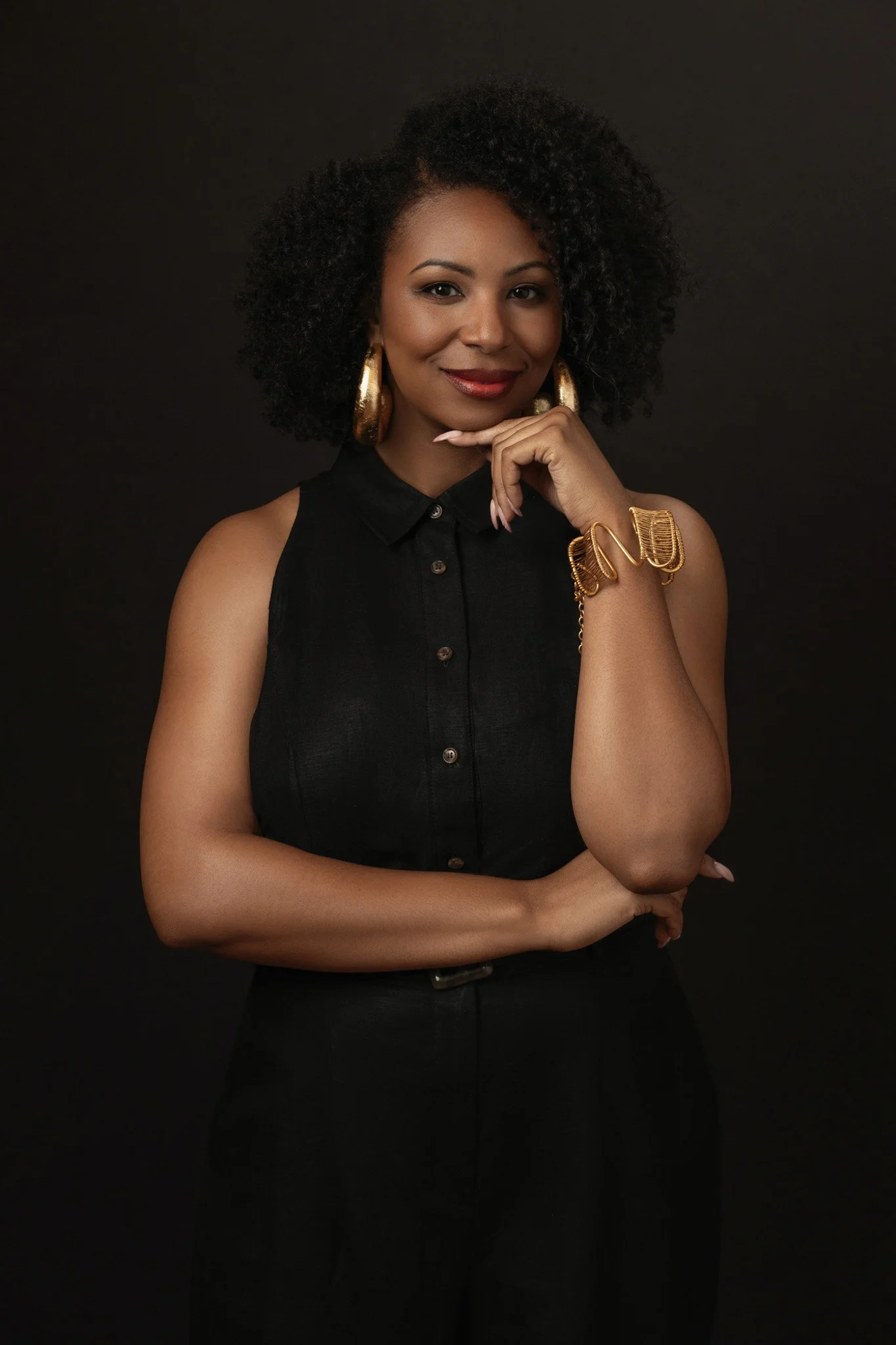 Black woman with natural curls and gold hoop earrings, hand to chin, against a black backdrop