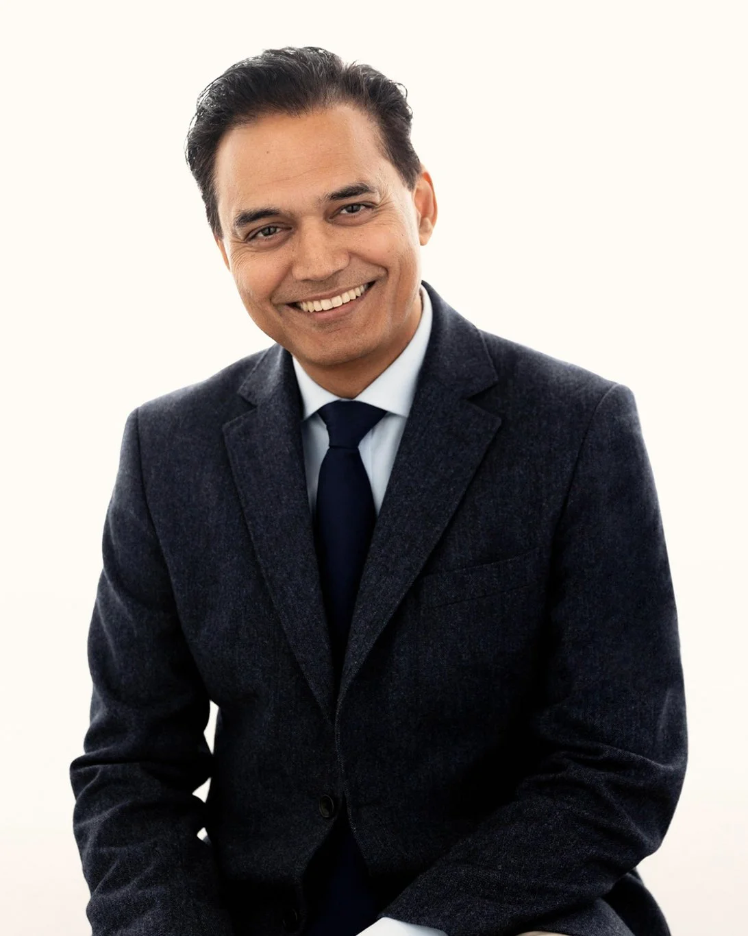 Smiling businessman in a charcoal blazer, light blue shirt and navy tie against a soft cream background