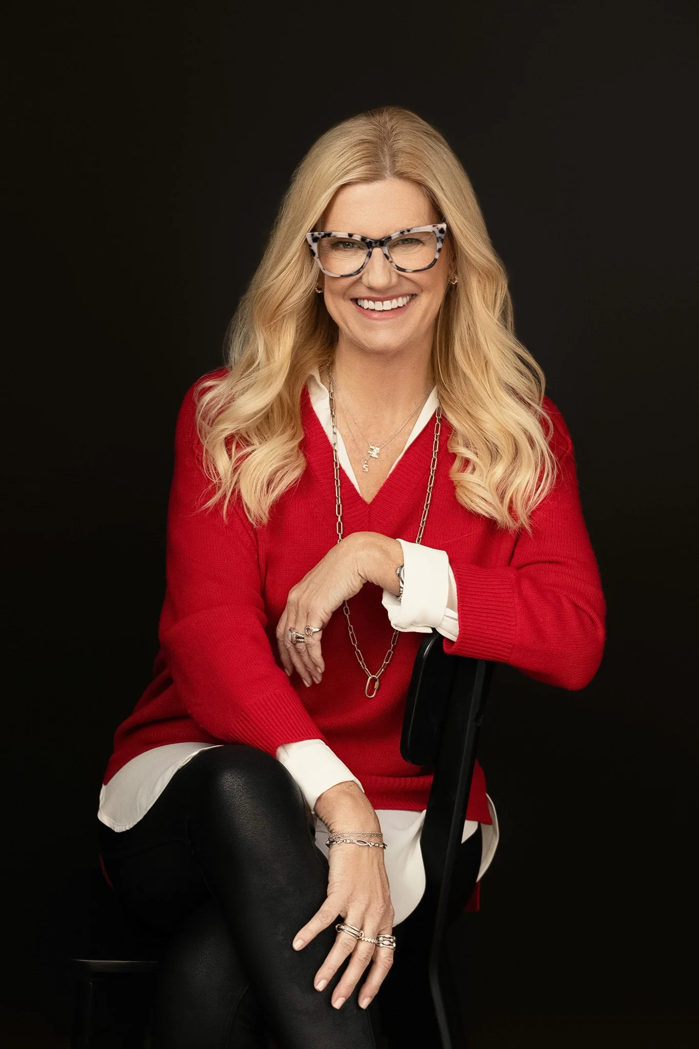 Blonde woman in tortoise glasses and a red sweater over white, seated on a stool against a black backdrop