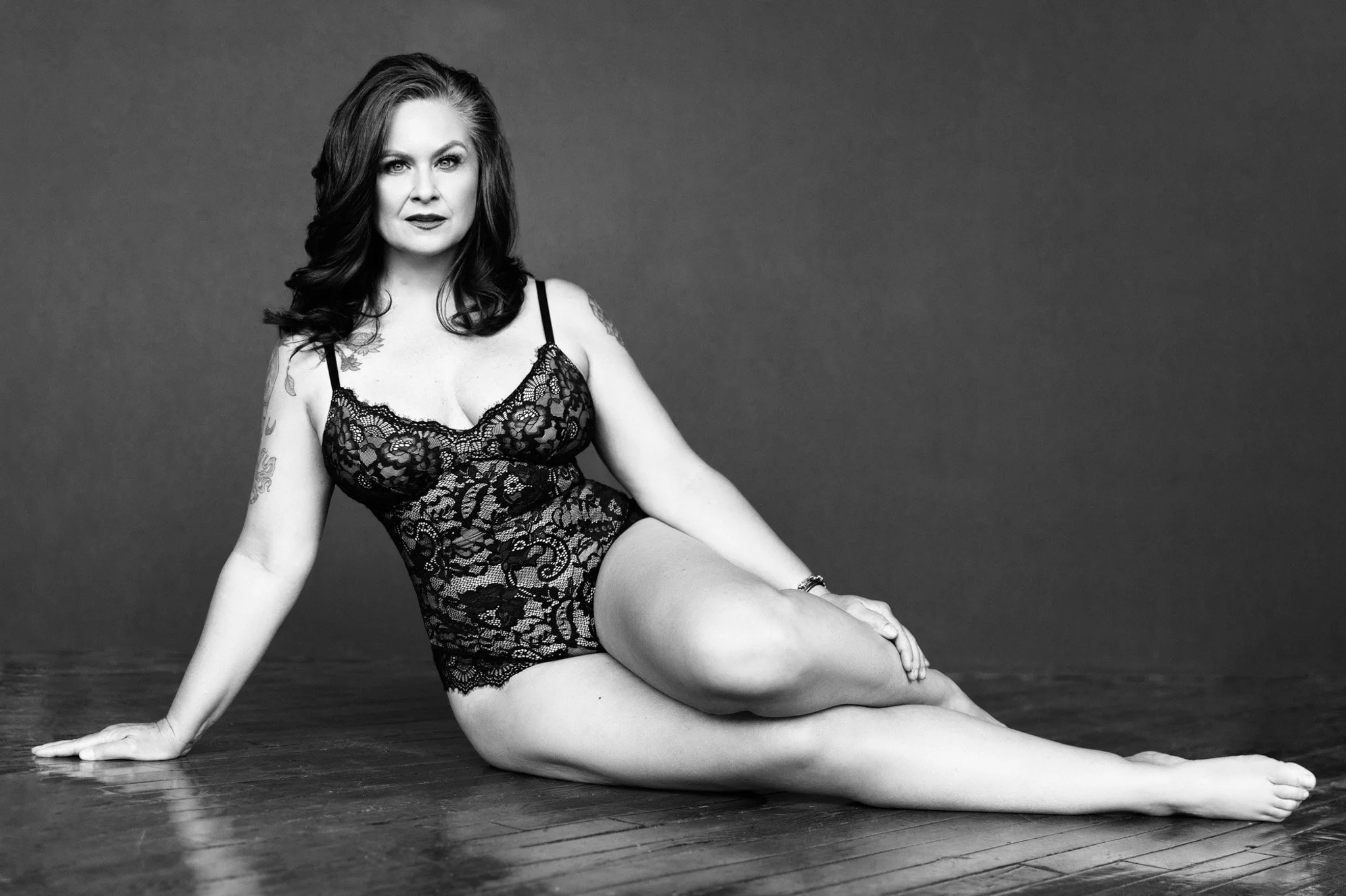 Curvy brunette woman reclining on the floor in a black lace bodysuit, confident and relaxed, black and white — Nashville boudoir portrait