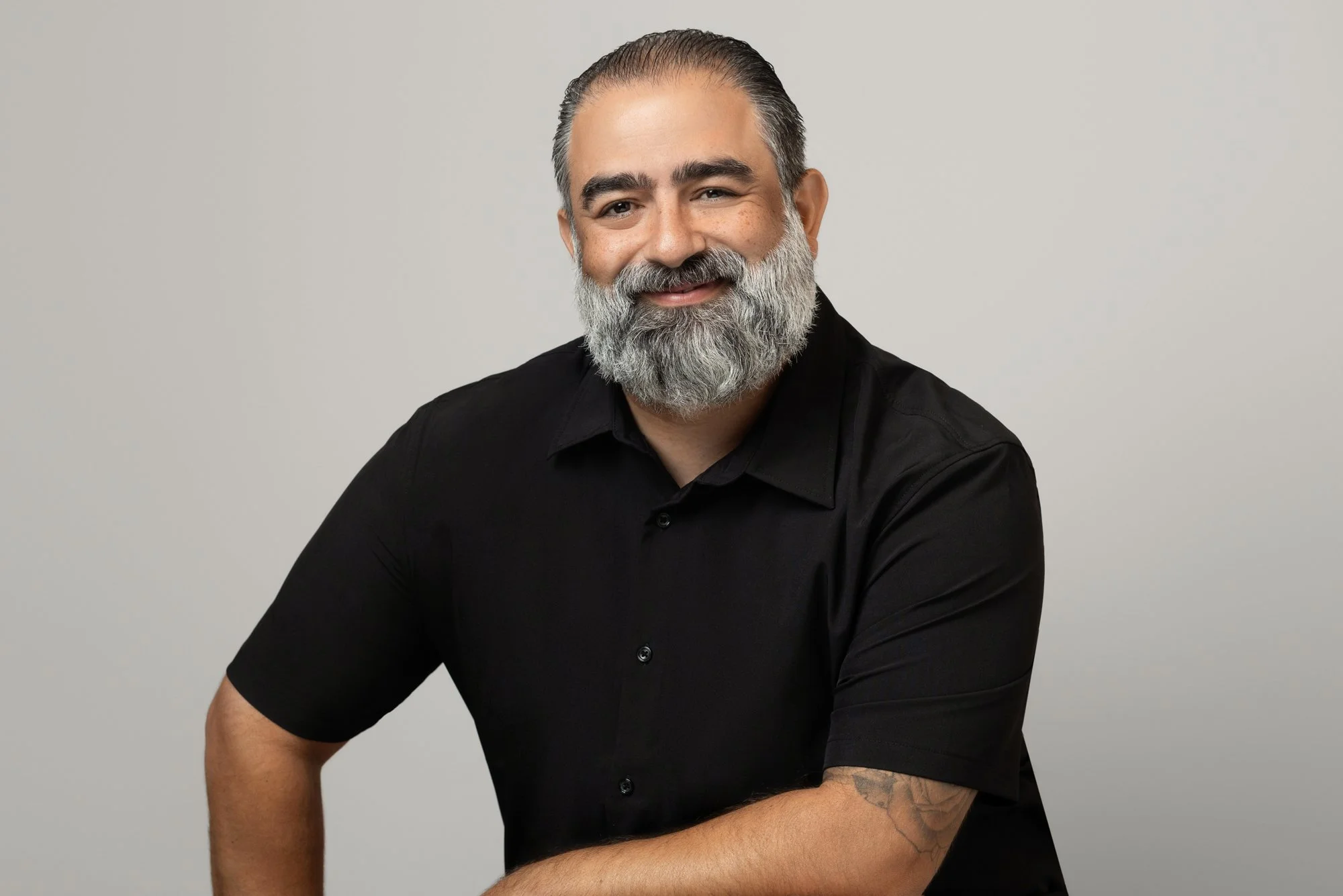 Corporate headshot of a man with a gray beard and slicked-back hair wearing a short-sleeve black button-up shirt, seated against a light gray background.