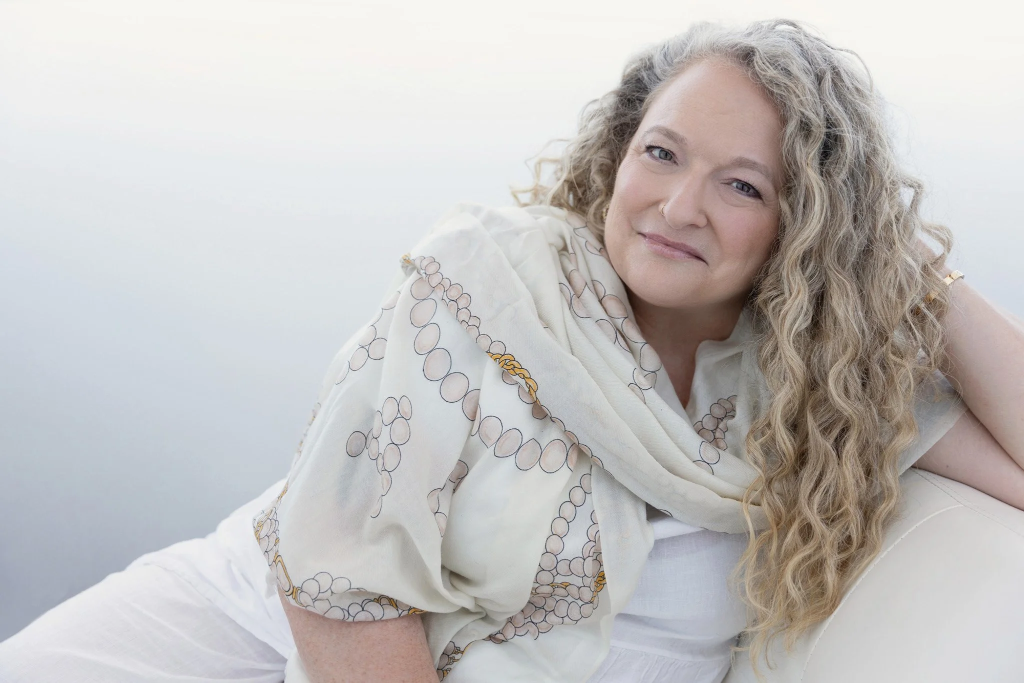 Woman with long curly hair in a cream scarf reclining against a soft light backdrop