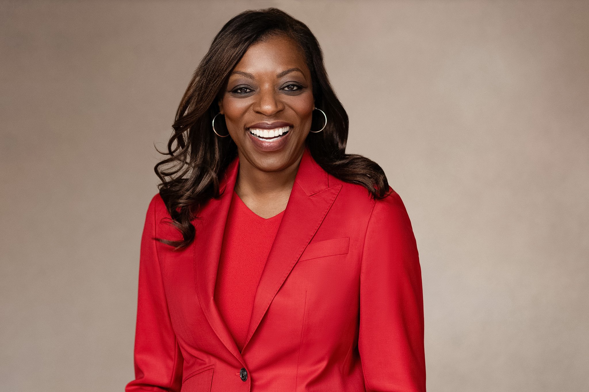 Black businesswoman in a red blazer with hoop earrings, laughing, against a warm taupe backdrop