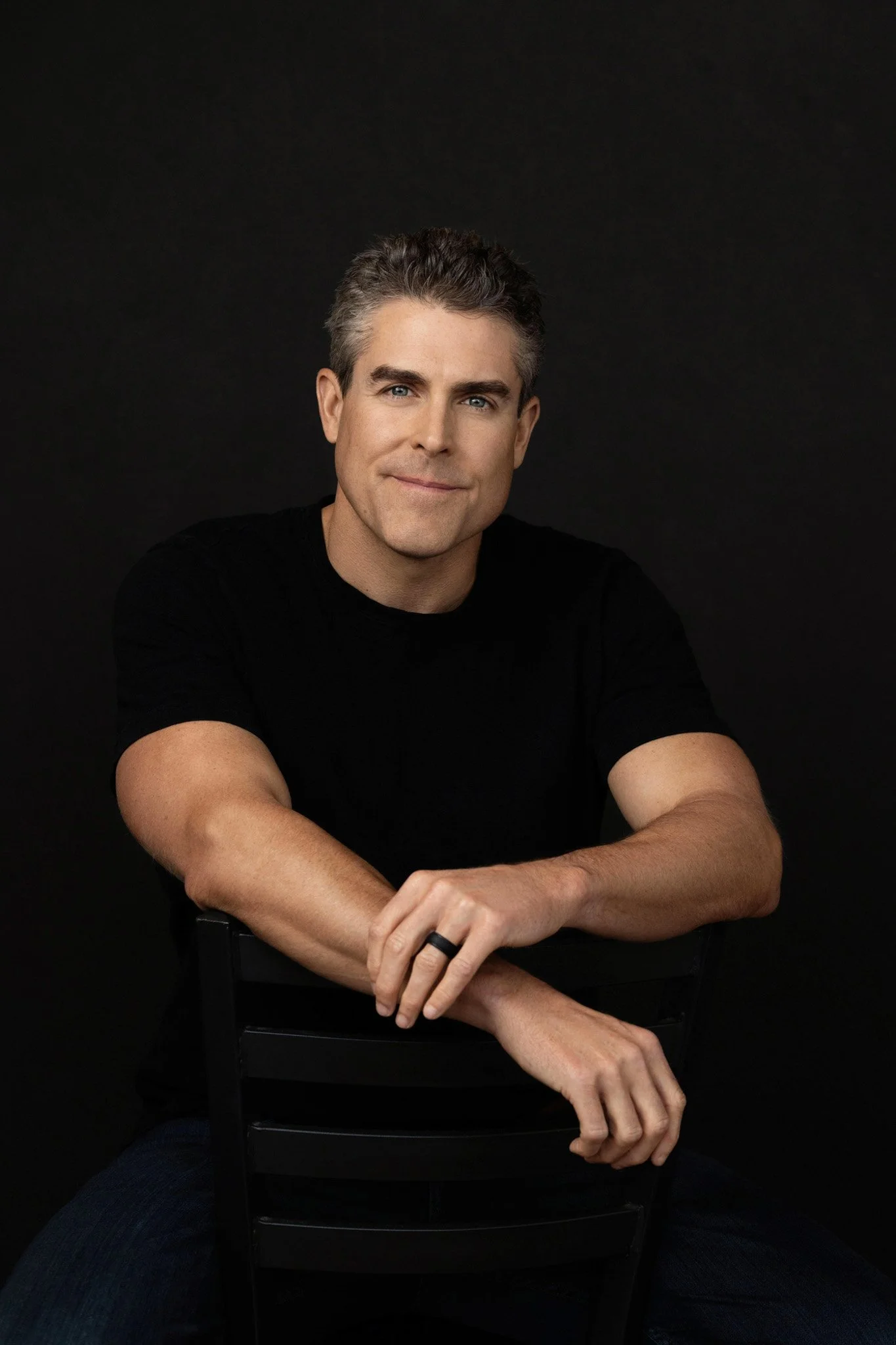 Man in a black t-shirt leaning on a chair back against a black backdrop