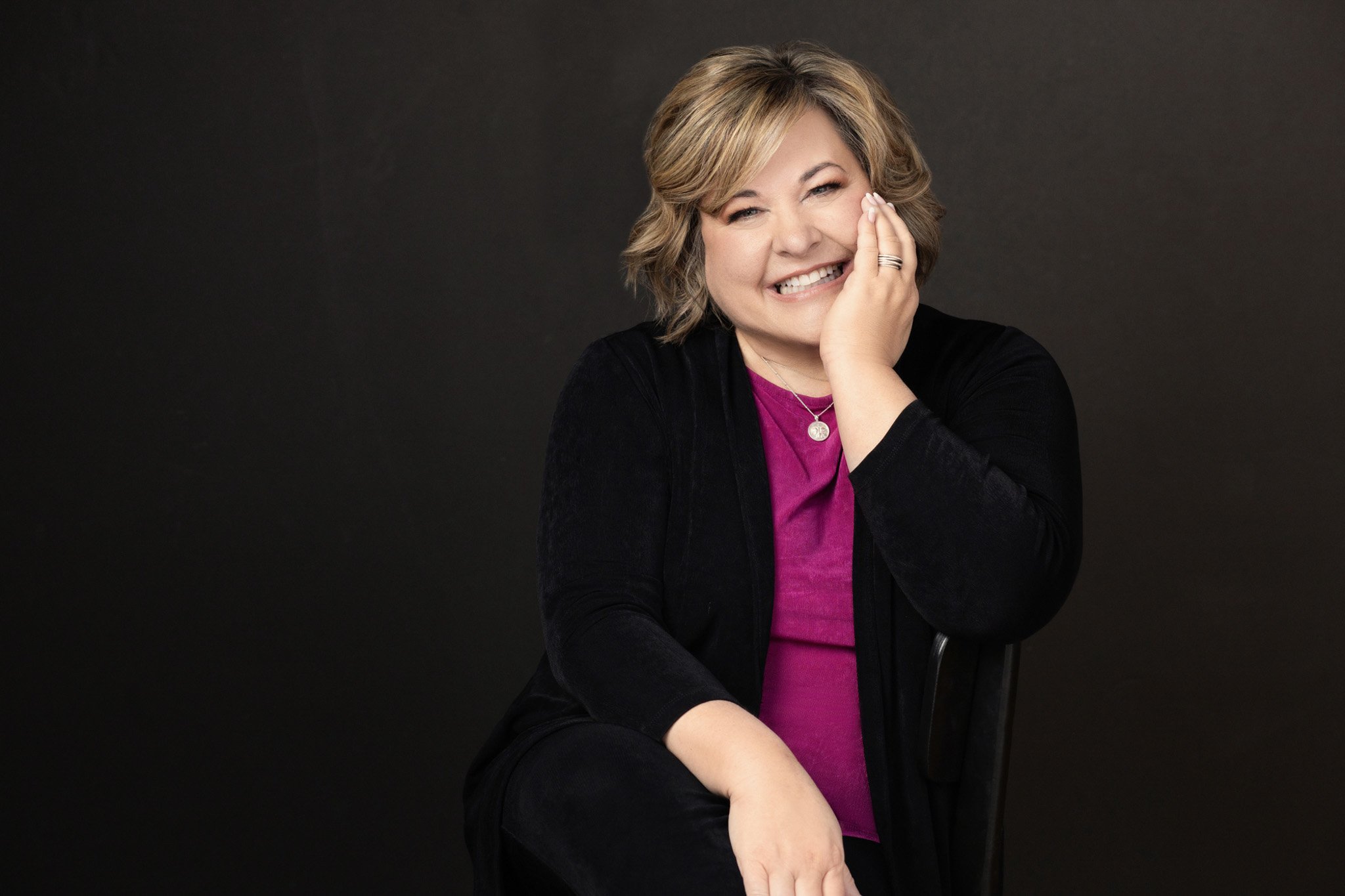 Woman in a black cardigan and magenta top laughing with hand to cheek against a black backdrop