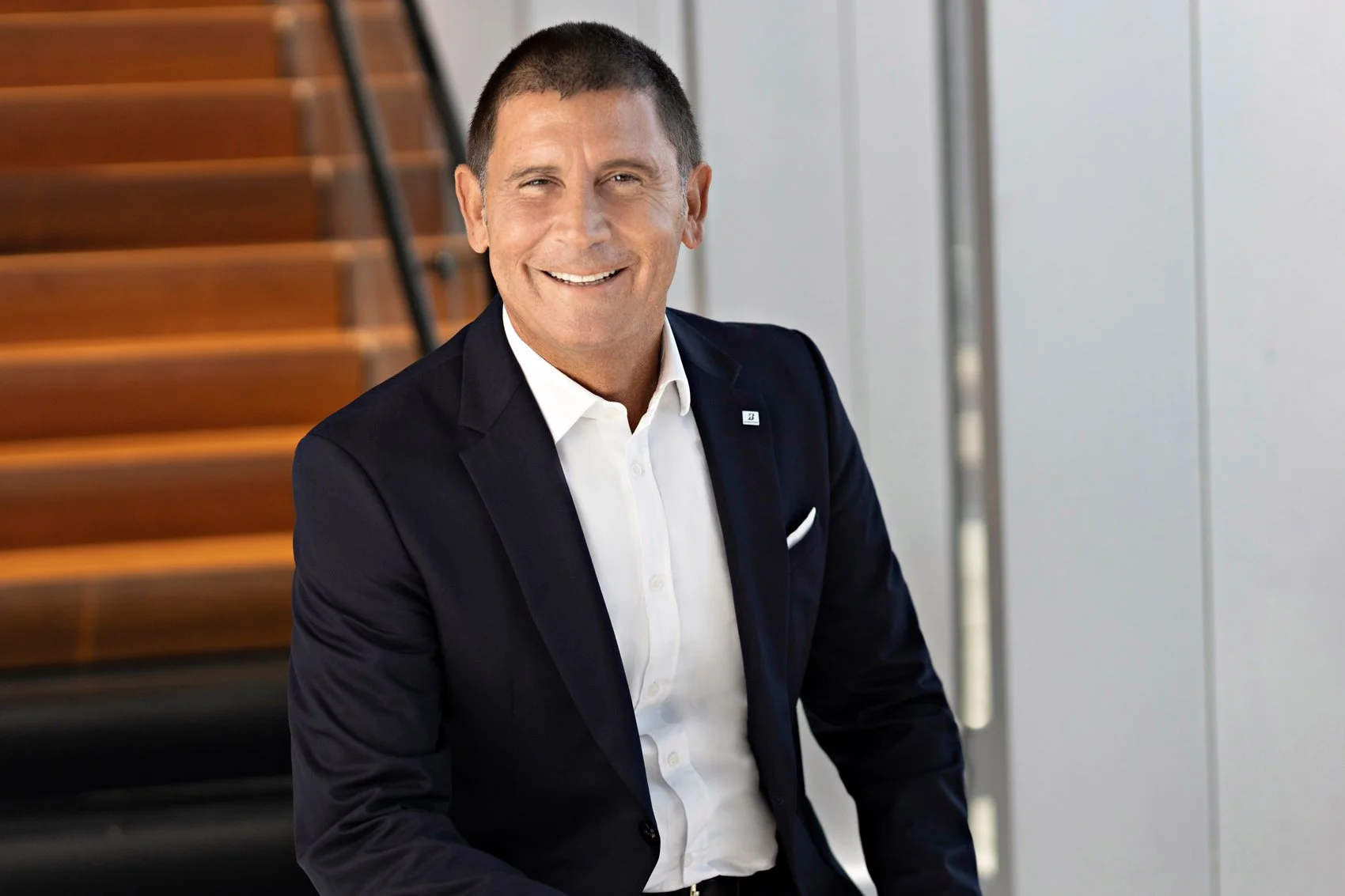 Executive headshot of a smiling man with short hair wearing a navy suit over a white shirt with a pocket square, beside a modern wooden staircase.