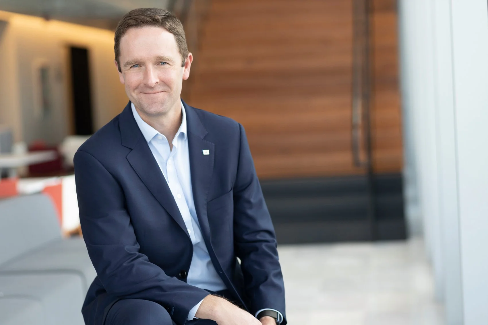 Executive headshot of a man wearing a navy suit over a light blue shirt, seated in a bright modern office with a wood-paneled stairway behind him.