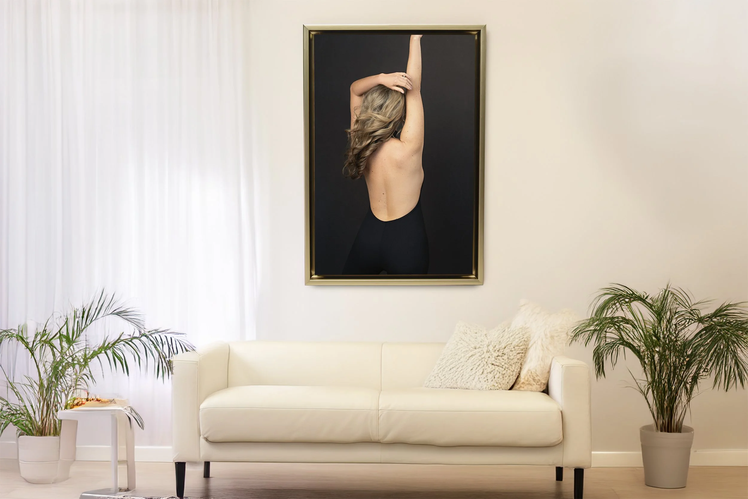 A large gold-framed vertical portrait hanging on a white wall above a cream-colored leather sofa. The artwork depicts the back of a woman with long blonde hair wearing a backless black outfit with her arms raised overhead.