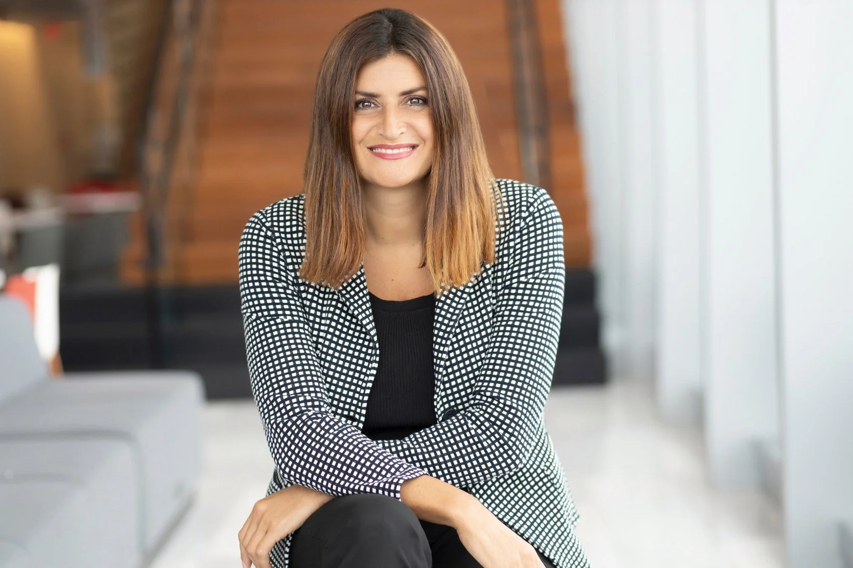 Corporate headshot of a smiling woman with shoulder-length brown hair wearing a black-and-white checkered blazer over a black top, in a modern office lobby.