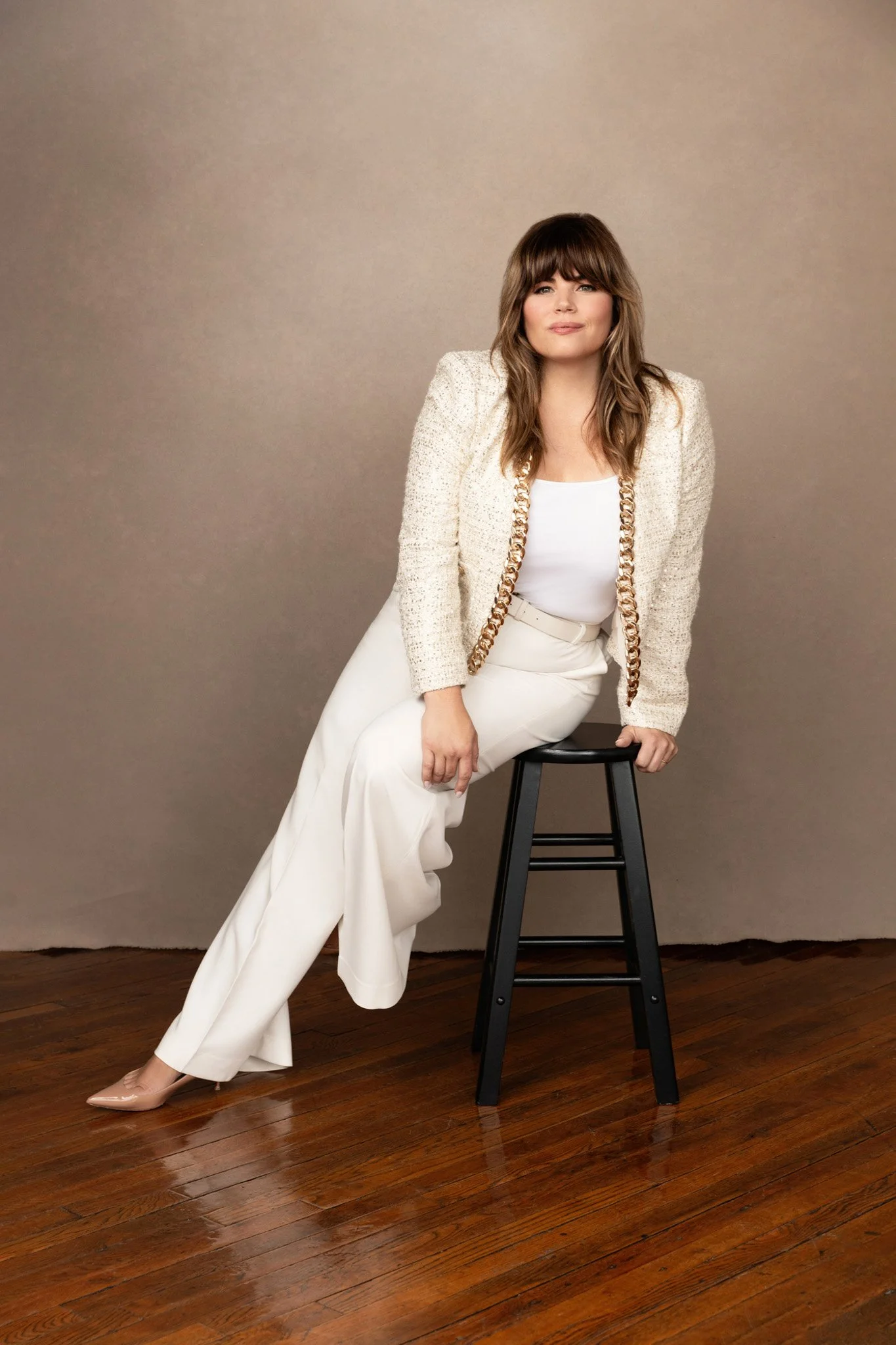 Brunette woman in a cream tweed jacket and white trousers seated full-length on a stool against a taupe backdrop