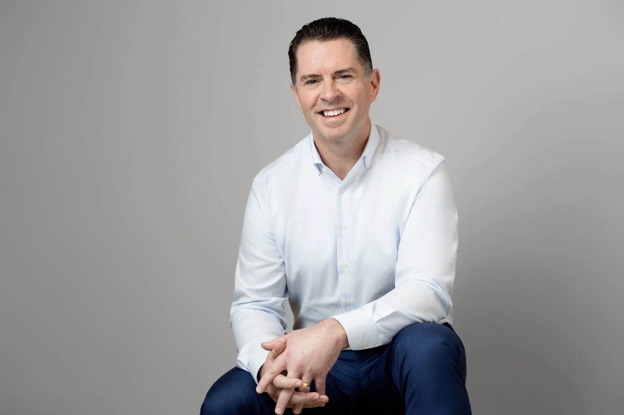 Business headshot of a smiling man with hands clasped wearing a light blue dress shirt and navy trousers, seated against a gray background.