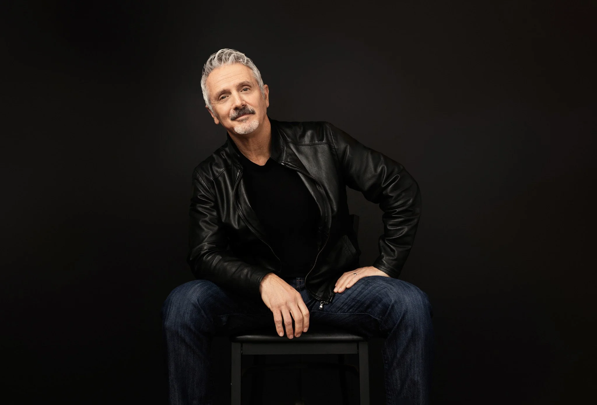 Man in a black leather jacket and jeans seated on a stool against a black backdrop