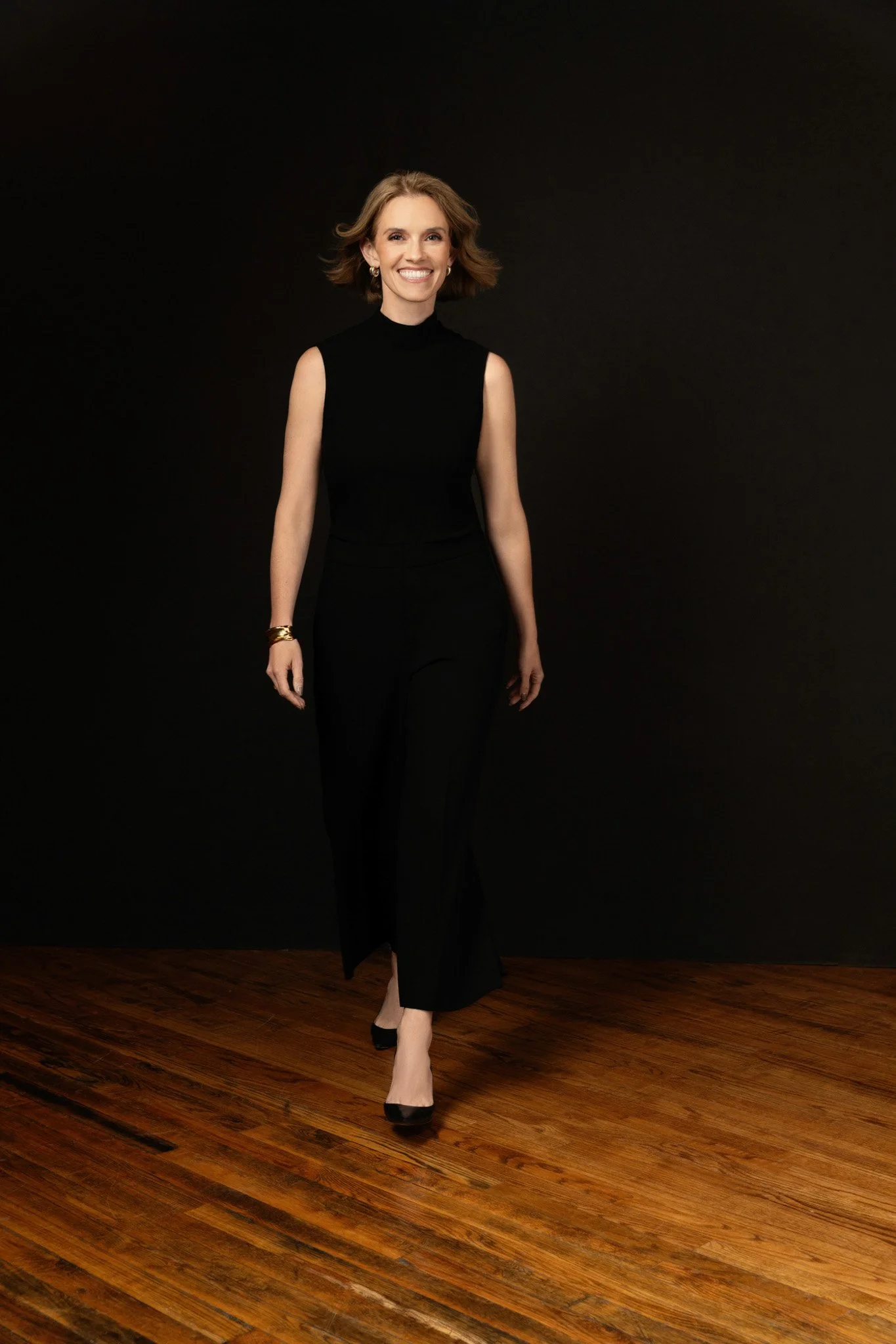 Blonde businesswoman in a black sleeveless jumpsuit walking across a wood floor against a black backdrop