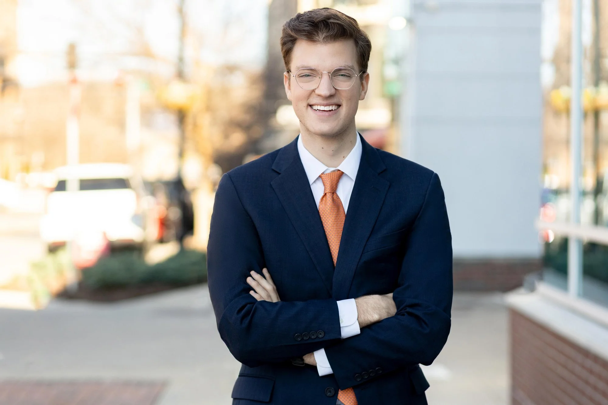 Outdoor business headshot of a young man with glasses and arms crossed wearing a navy suit with an orange tie, on a blurred city street.