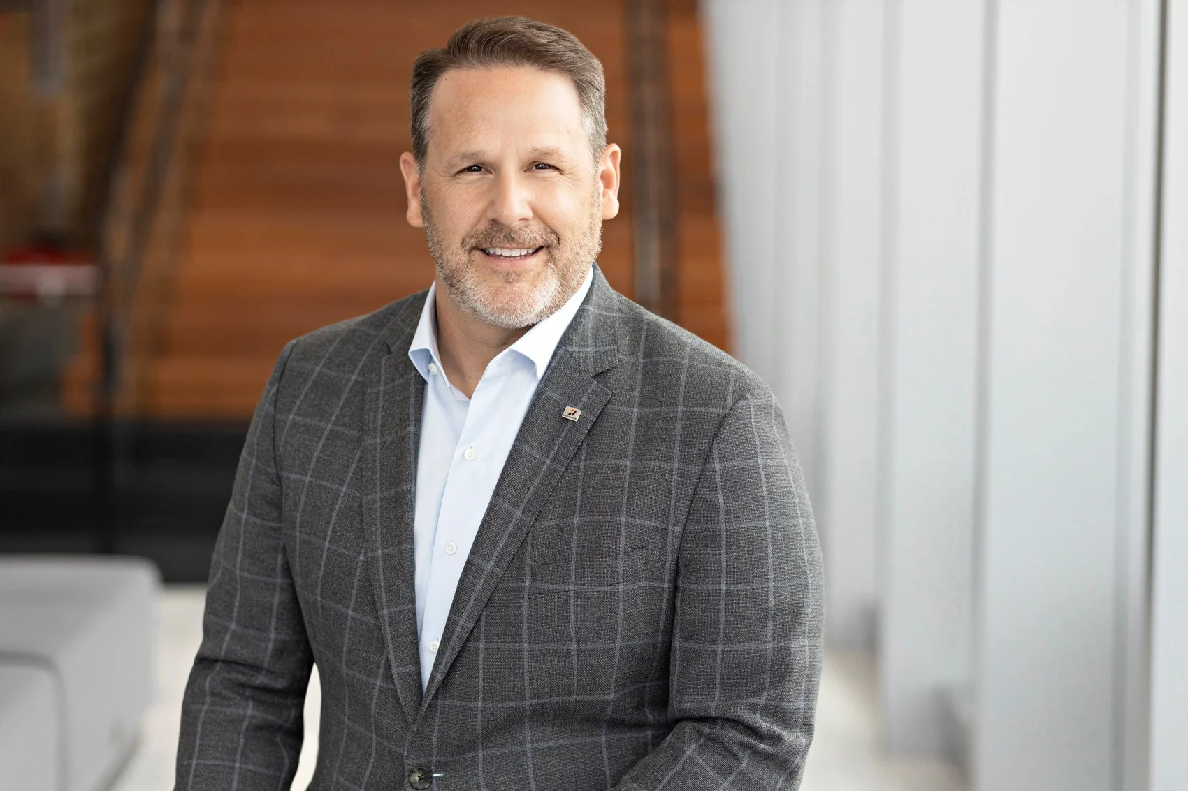 Executive headshot of a smiling man with short gray hair and a beard wearing a gray windowpane-check blazer over a light blue shirt, in a modern office space.