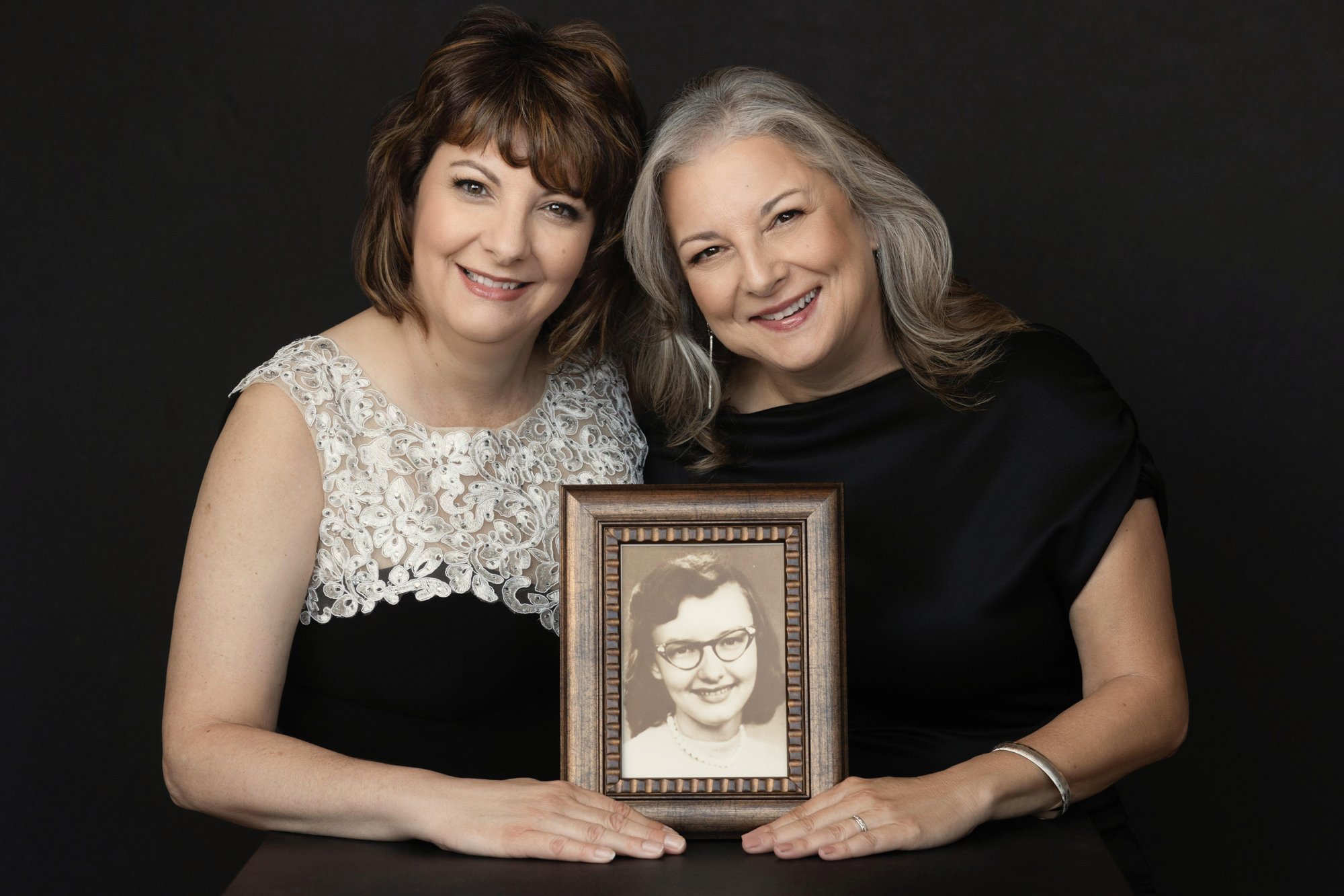 Two women smiling while holding a framed vintage portrait of an older relative, honoring family history and generational bonds through family photography.