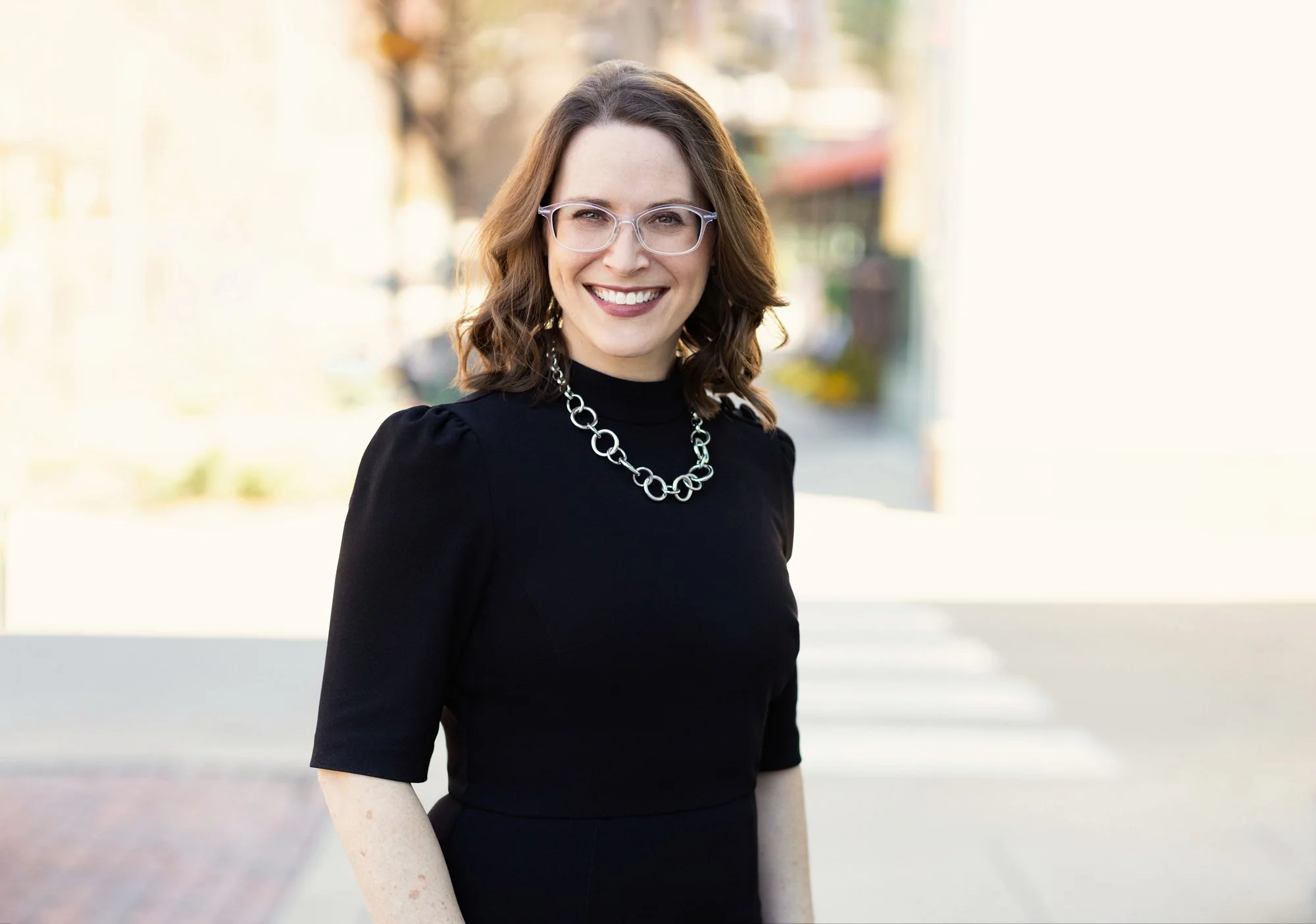 Outdoor business headshot of a smiling woman wearing glasses, a black dress, and a chunky silver necklace, with a blurred downtown street behind her.