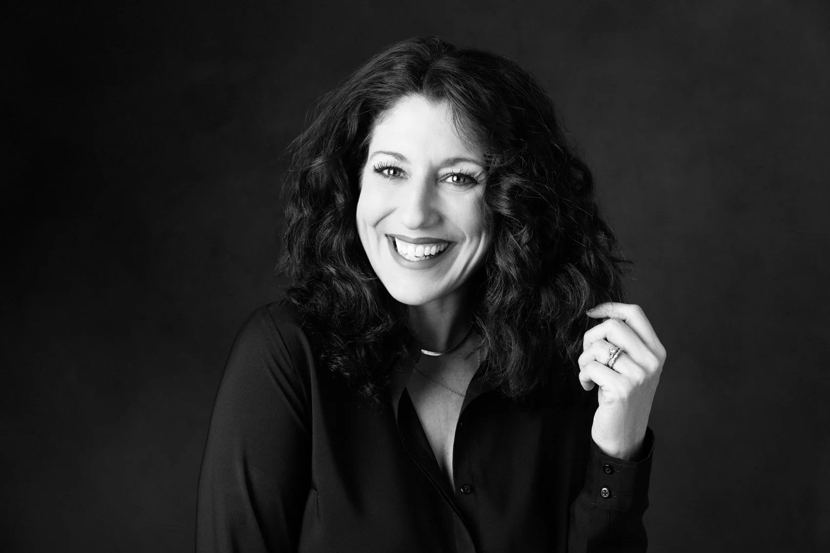 Black-and-white portrait of Nashville boudoir and portrait photographer Judith Hill, laughing with dark curly hair and a black blouse against a dark background