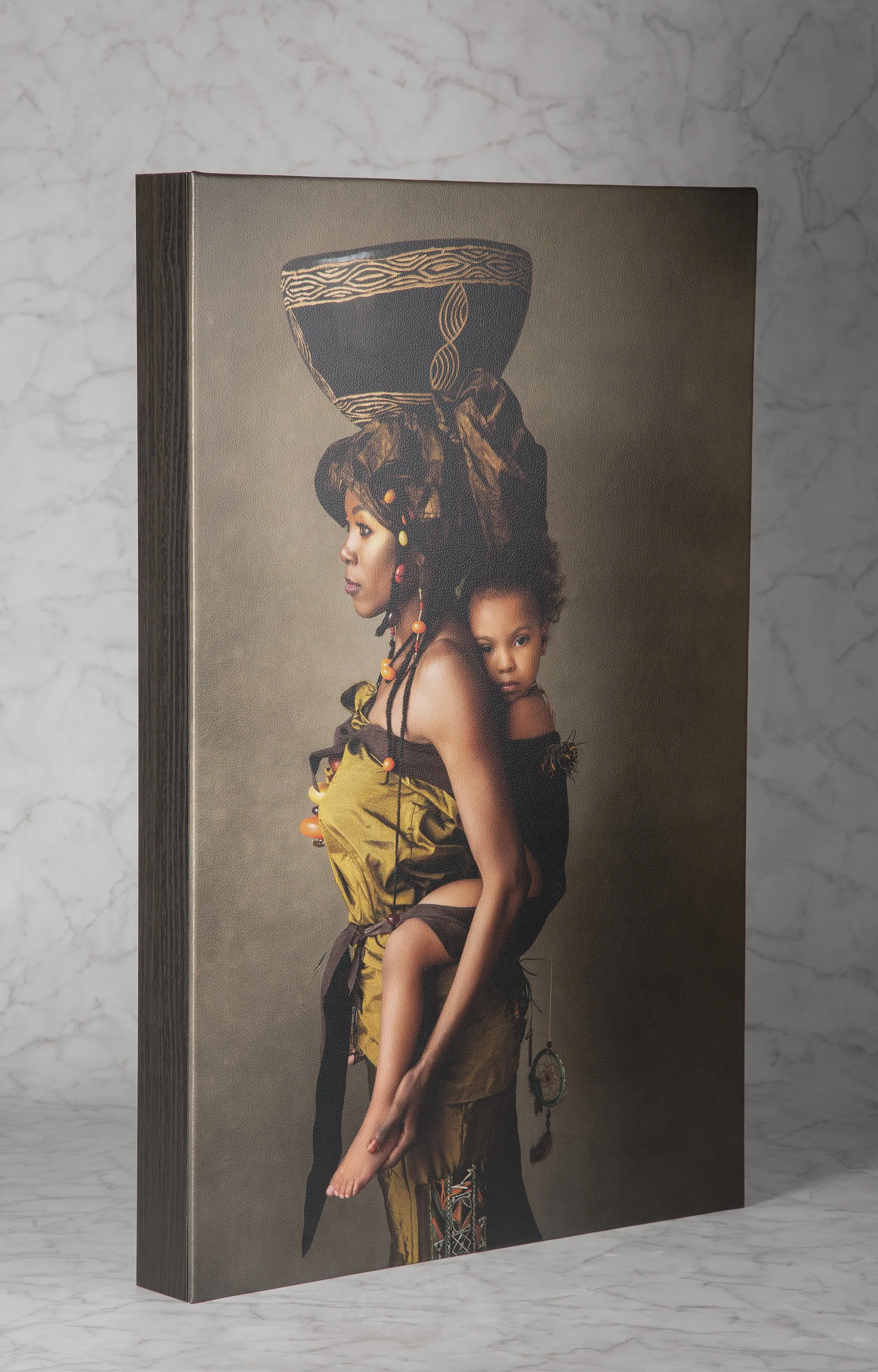 A thick wooden block canvas print standing upright on a white marble surface. The artwork depicts a profile view of a Black woman in traditional golden-brown clothing carrying a young child on her back.