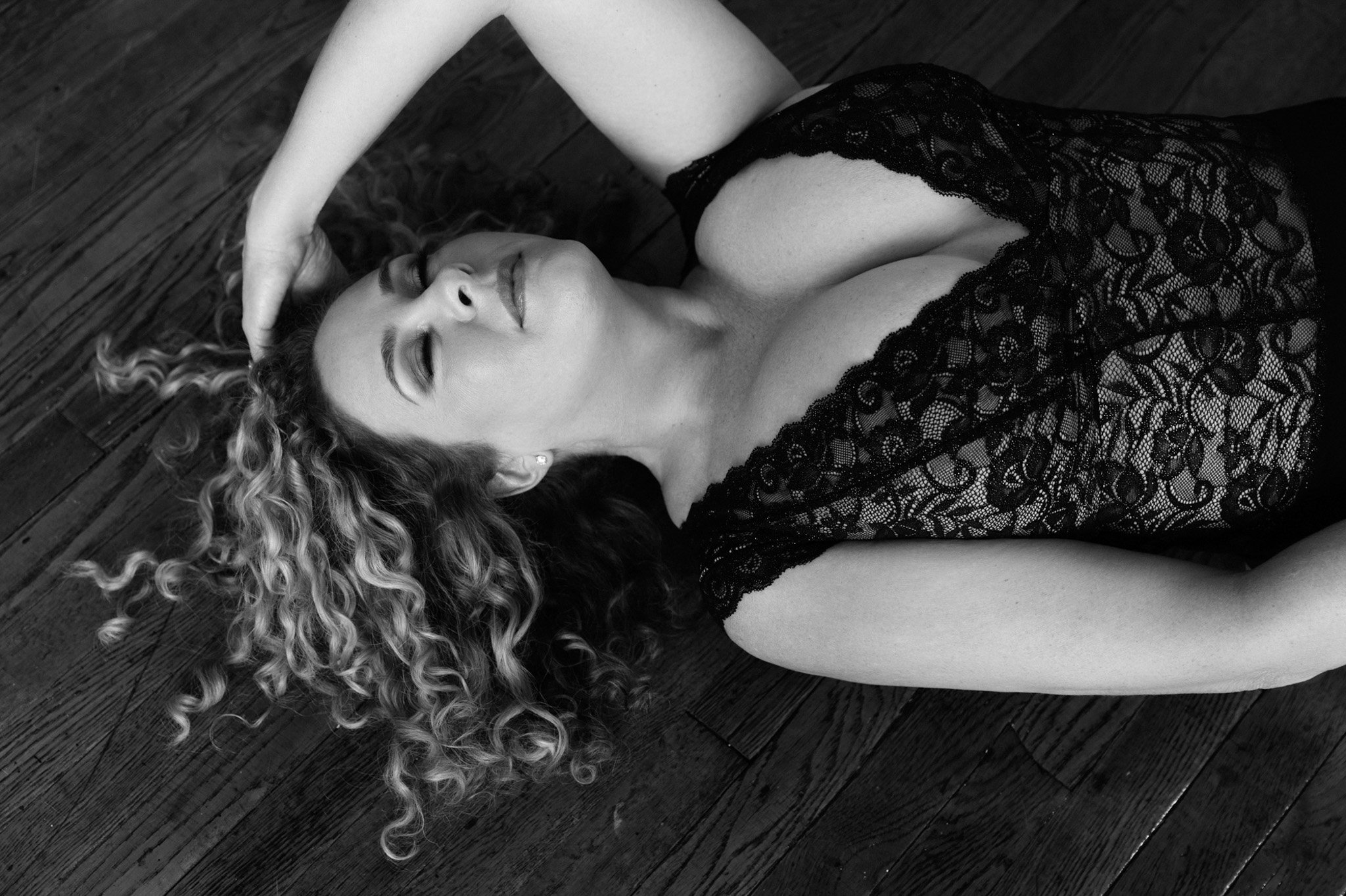Curly-haired blonde woman reclining on a hardwood floor in a black lace bodysuit, eyes closed, black and white — Nashville boudoir