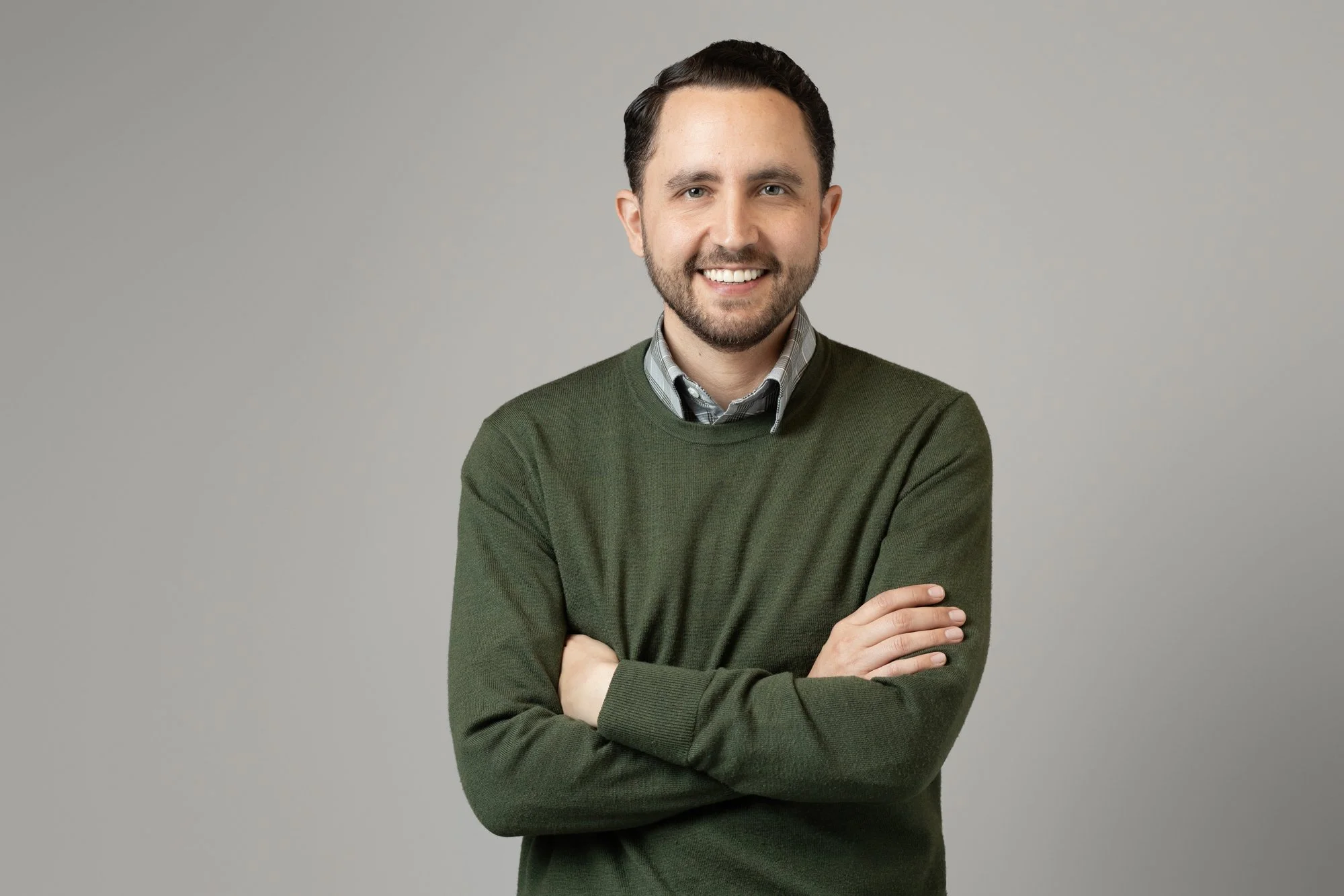 Business headshot of a smiling man with arms crossed wearing a green crewneck sweater over a plaid collared shirt, on a gray studio backdrop.