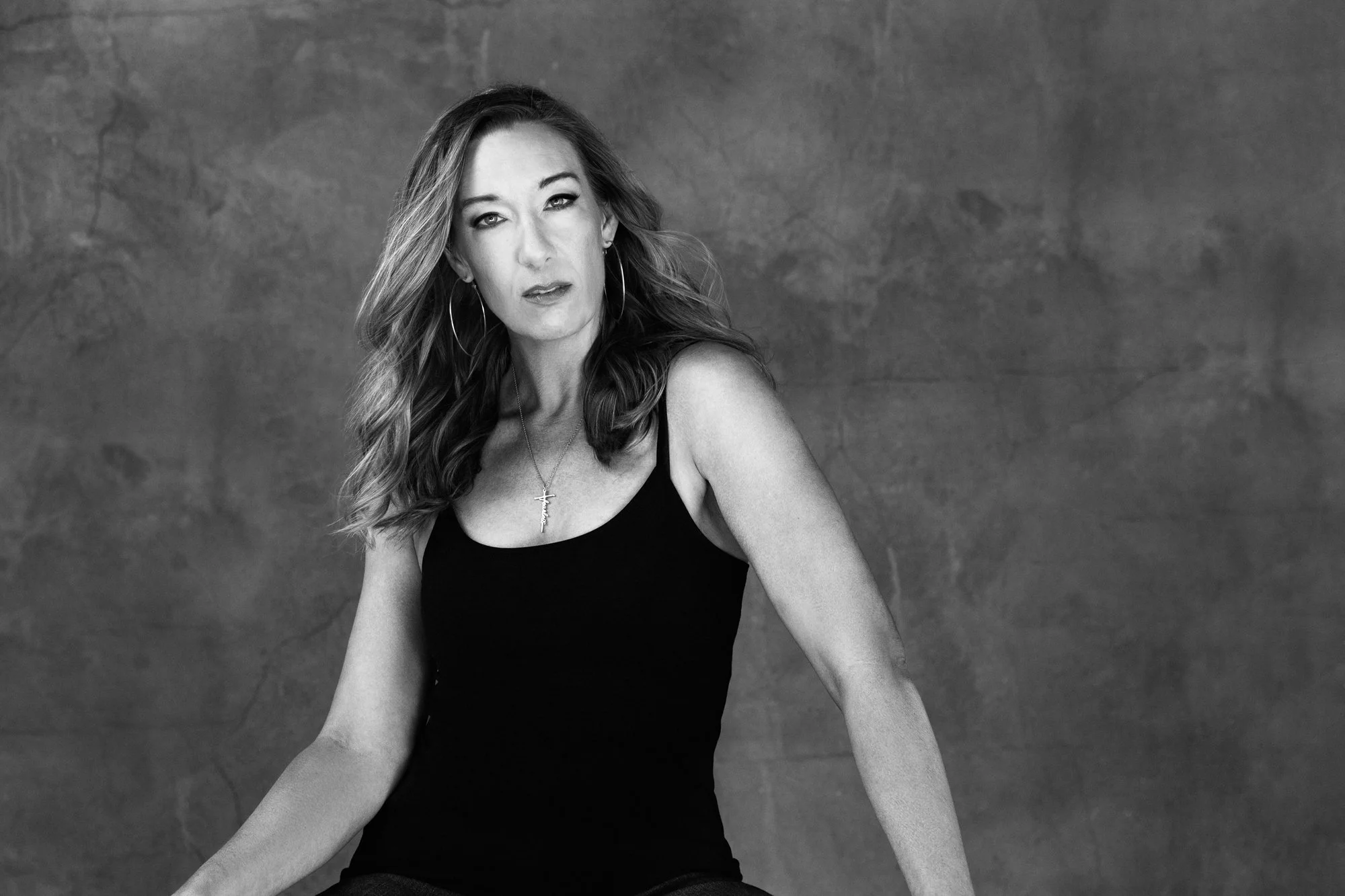 Woman with long wavy hair in a black tank top looks confidently toward the camera against a simple studio backdrop. The portrait highlights strength and presence often celebrated in women’s portrait photography.