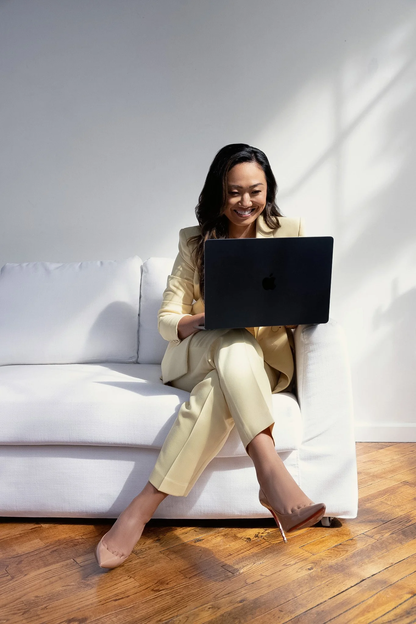 Woman in a pale yellow suit working on a laptop on a white sofa in natural light