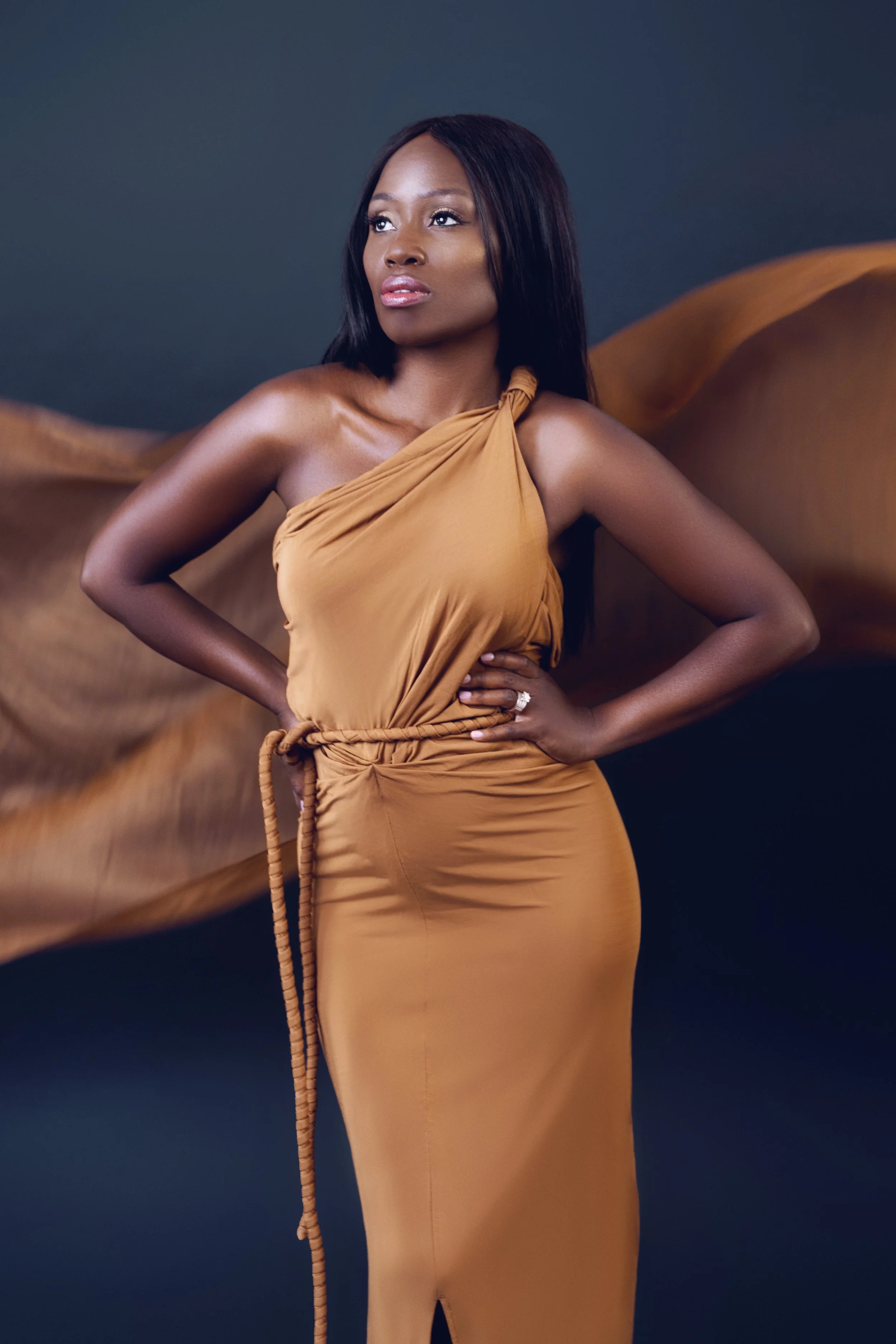 Dark-haired black woman in a flowing one-shoulder caramel gown with hands on her hips and fabric streaming behind her, Nashville studio portrait by Judith Hill
