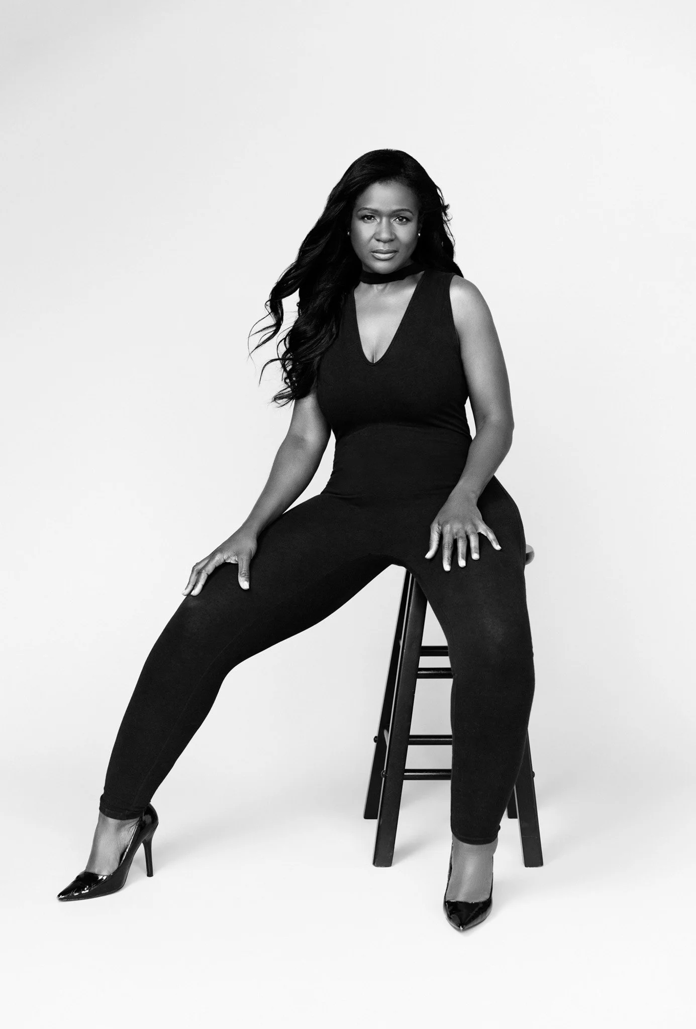 Long-haired woman in a black sleeveless jumpsuit and heels seated on a stool, igh-key black-and-white Nashville studio portrait by Judith Hill