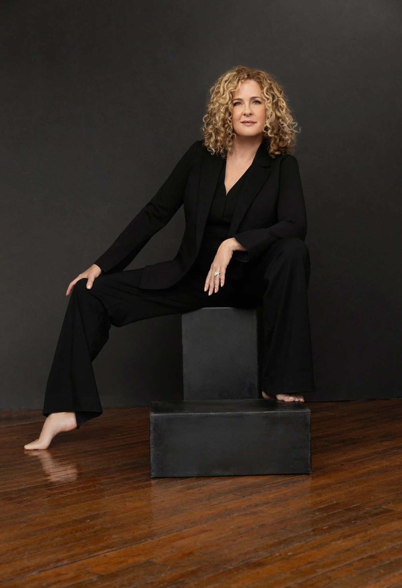 Woman in an all-black suit seated barefoot on gray blocks against a gray backdrop