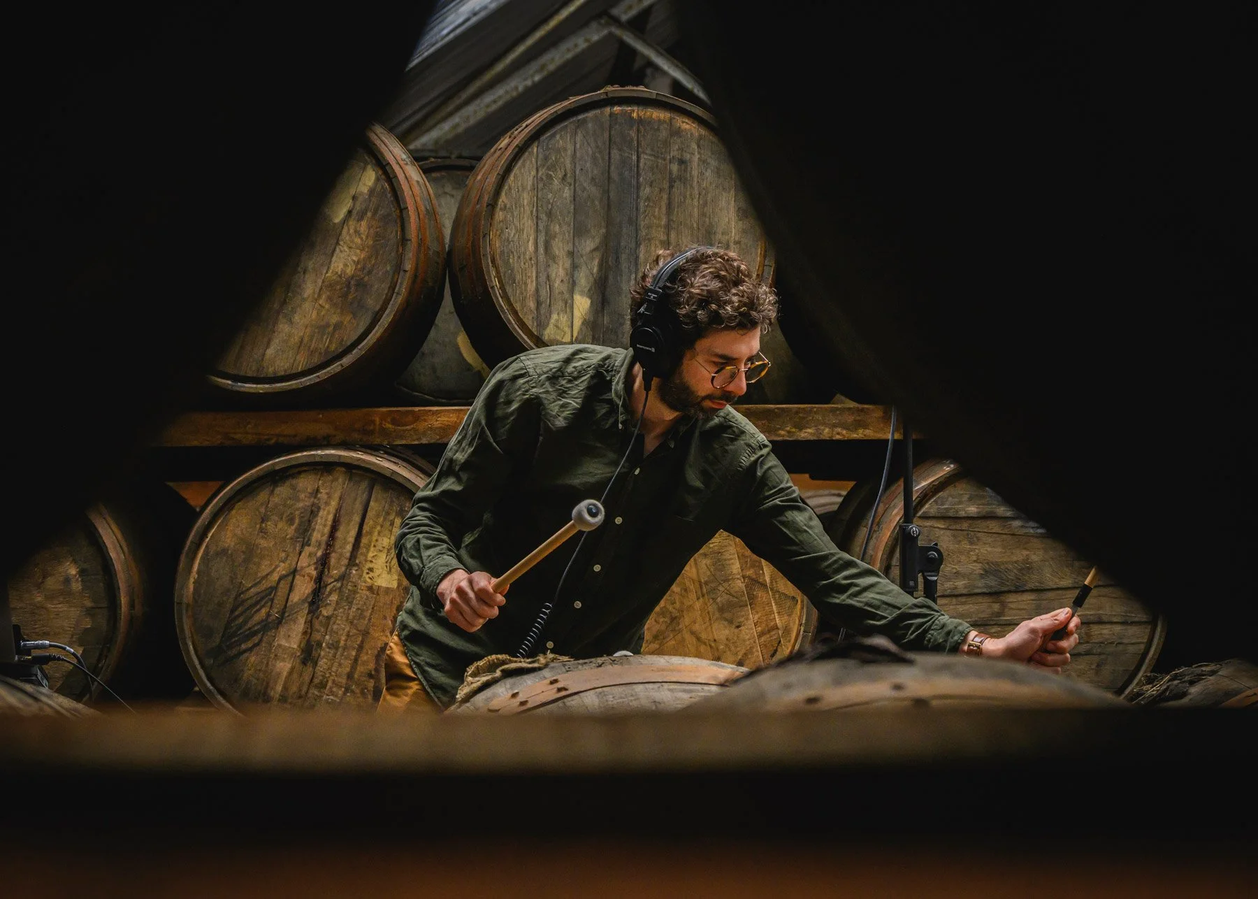Man with curly hair, glasses, and headphones playing a drum with a stick in a cellar or wine cellar with wooden barrels in the background.