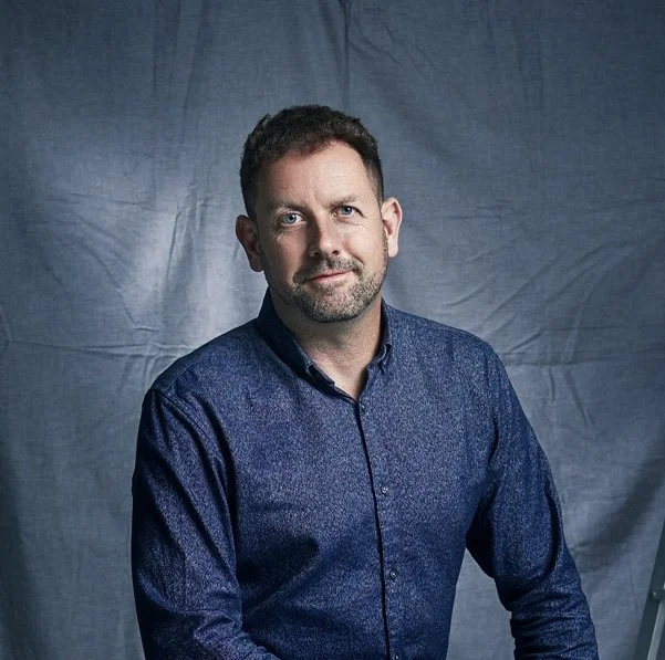 A man with short dark hair and a beard wearing a dark blue shirt, sitting in front of a gray backdrop.